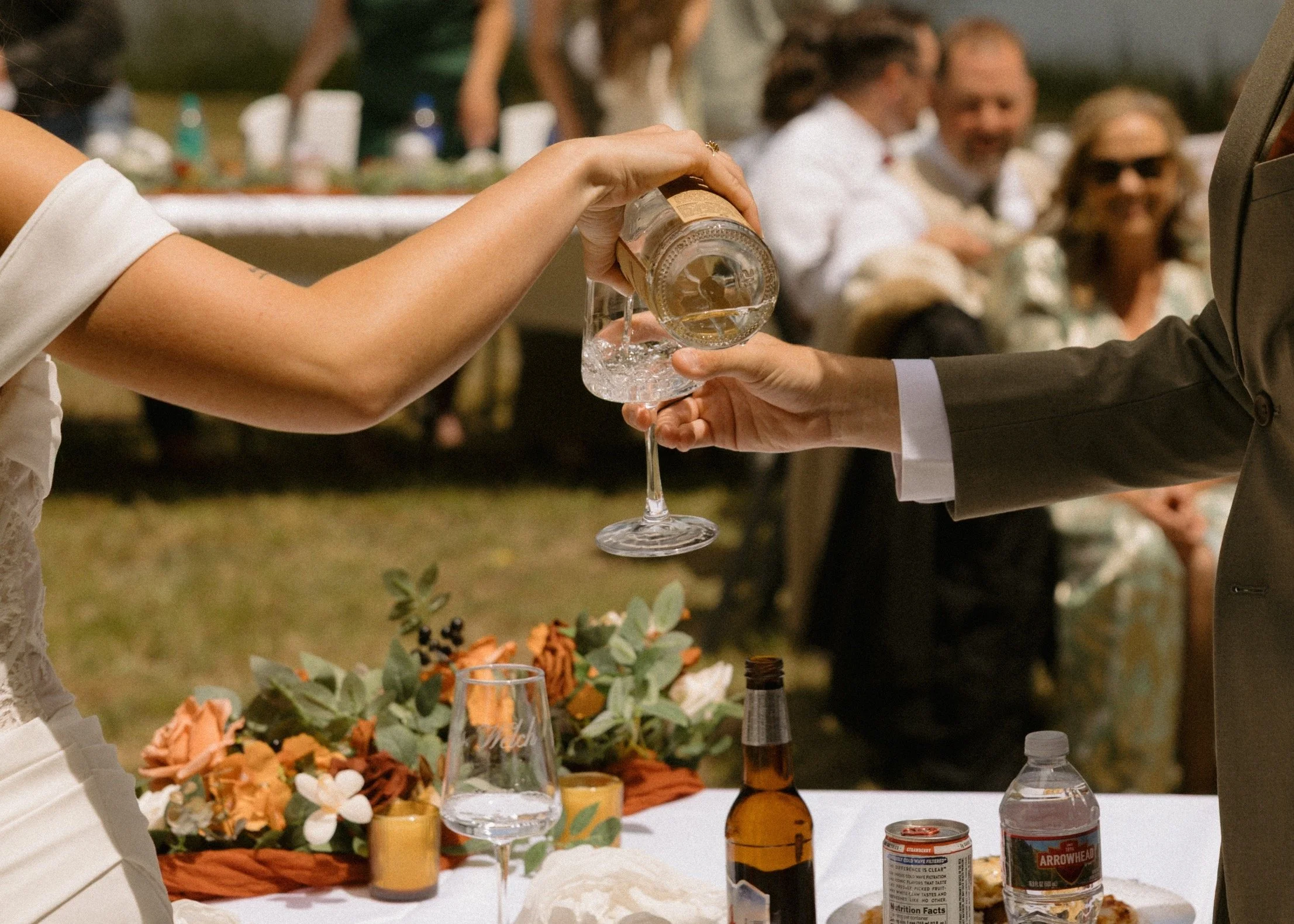 A wedding reception scene with a person pouring a drink into a glass held by a man in a suit. The table is decorated with flowers, candles, and bottles, and in the background, guests are seated outdoors, smiling and chatting.