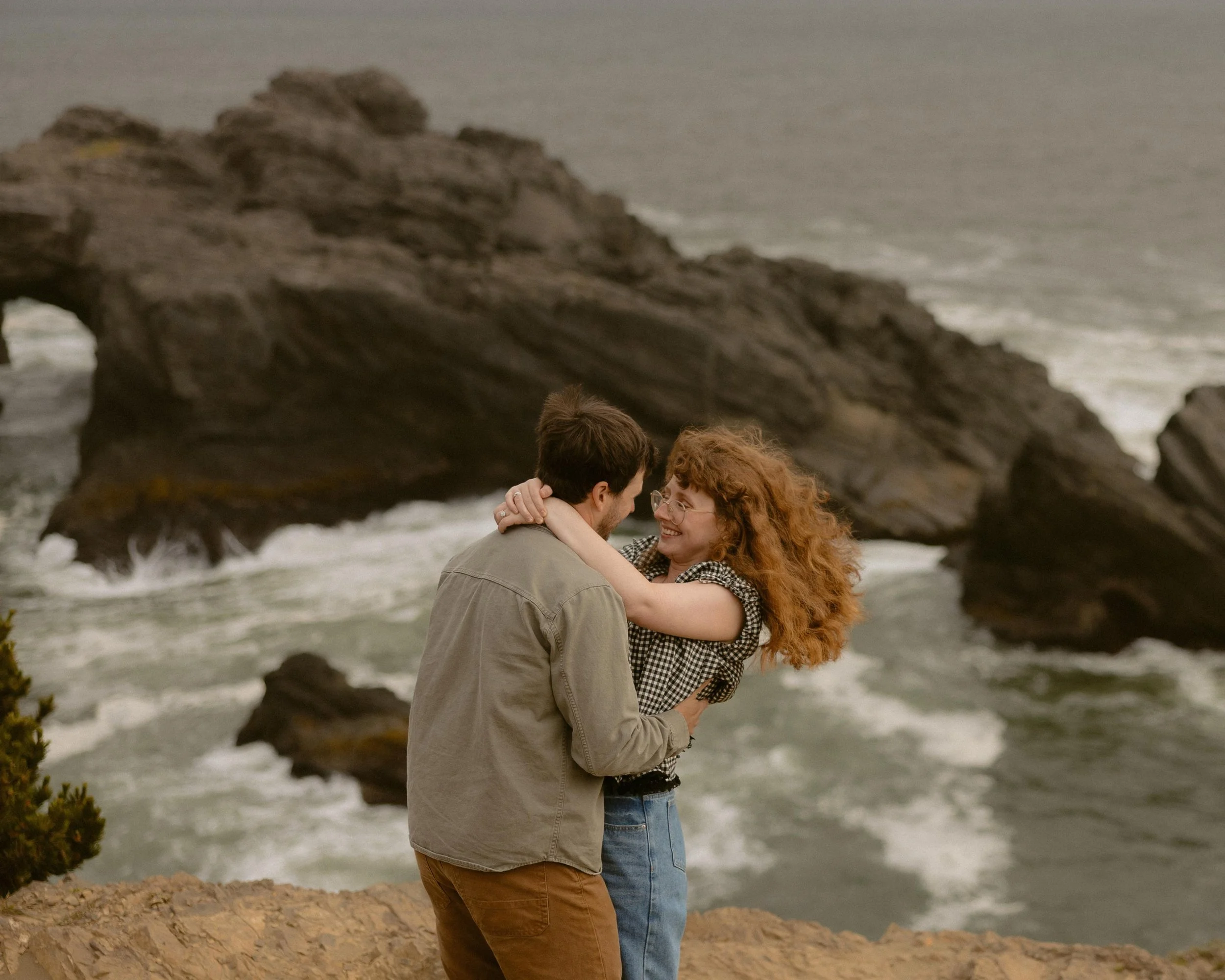 A couple kissing on a rocky beach with large rock formations in the background.