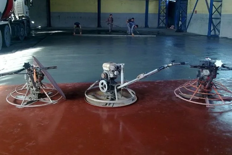 Three concrete trowels with motorized handles smoothing freshly poured concrete floor in a large industrial space.