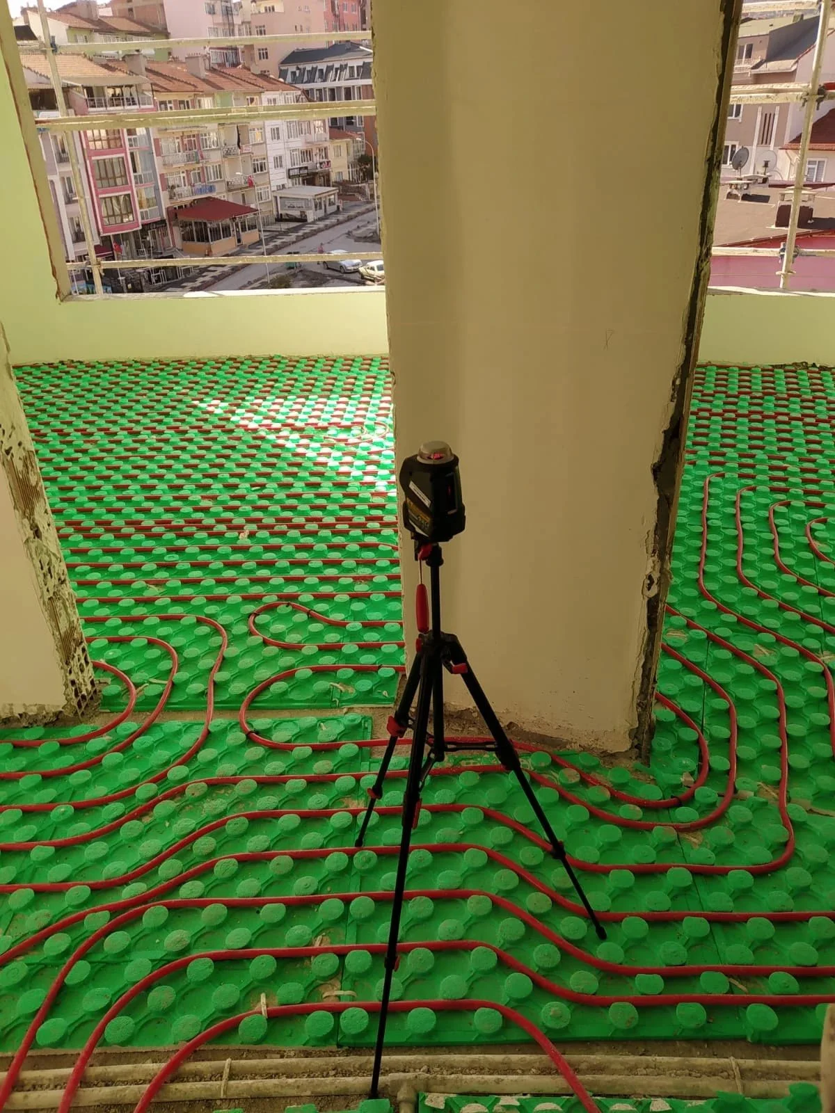 Room under construction with green insulation panels and red pipes on the floor, a tripod with a laser level in the center, and city buildings visible through large windows.