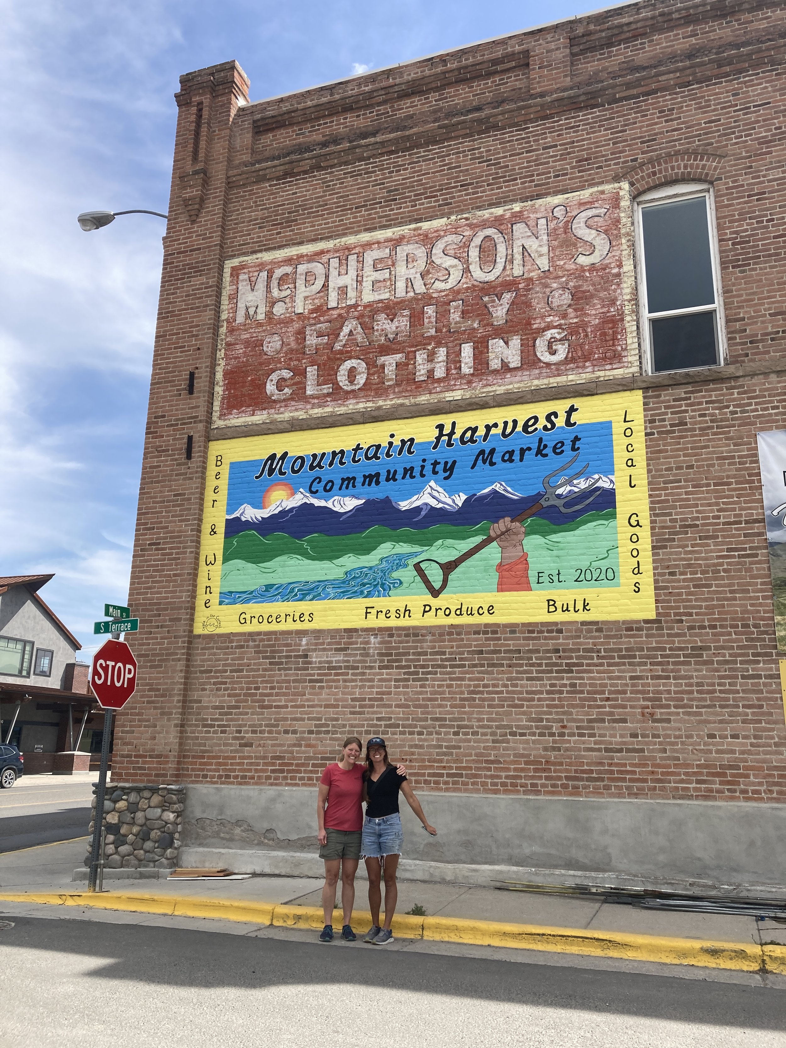 Two women stand on a sidewalk in front of a large brick building with a painted mural advertising Mountain Harvest Community Market, established in 2020, offering groceries, fresh produce, and bulk goods, with a backdrop of mountains and a river. The