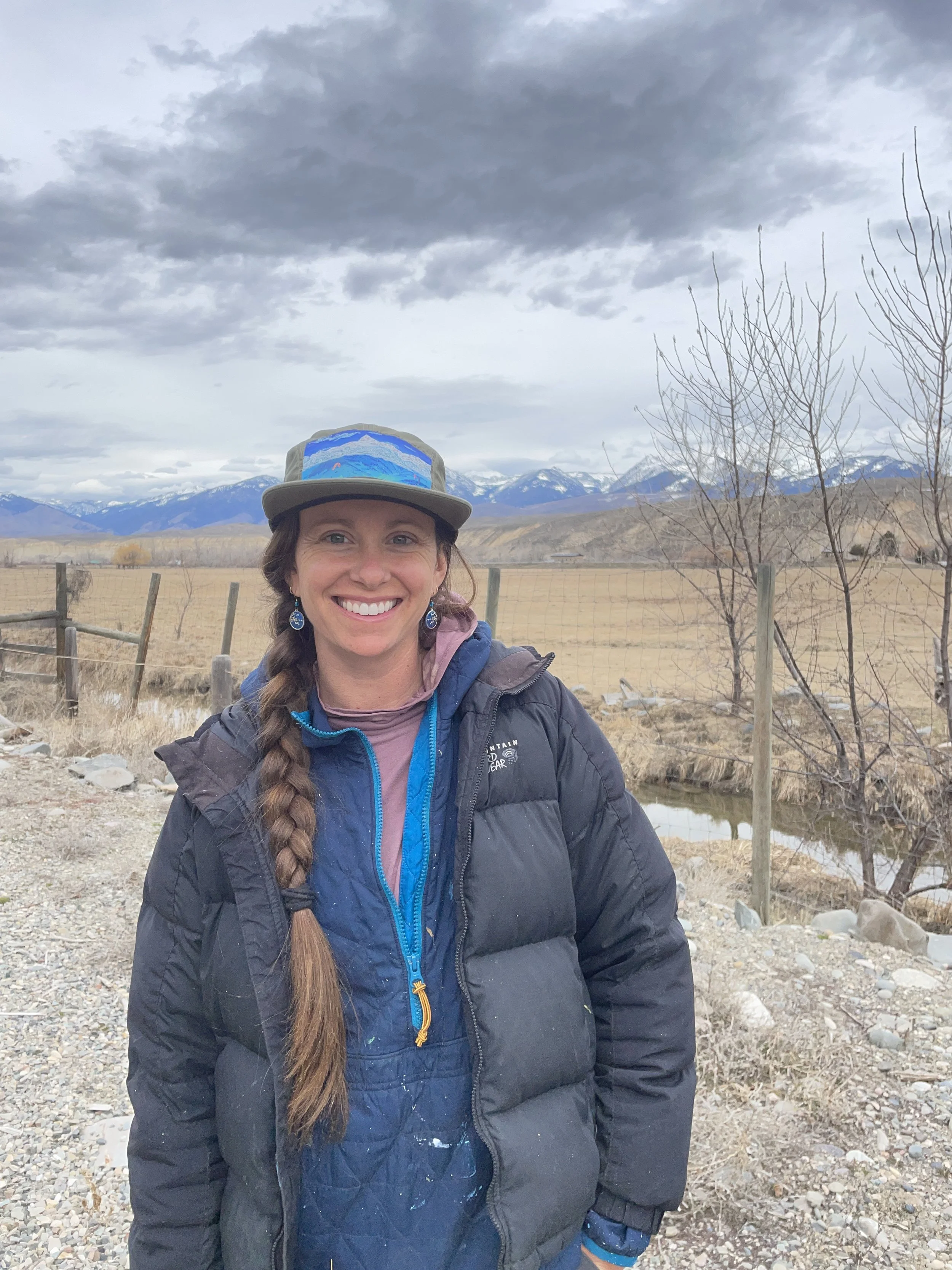 Woman with long braided hair smiling outdoors near a fence, with mountains and cloudy sky in the background.