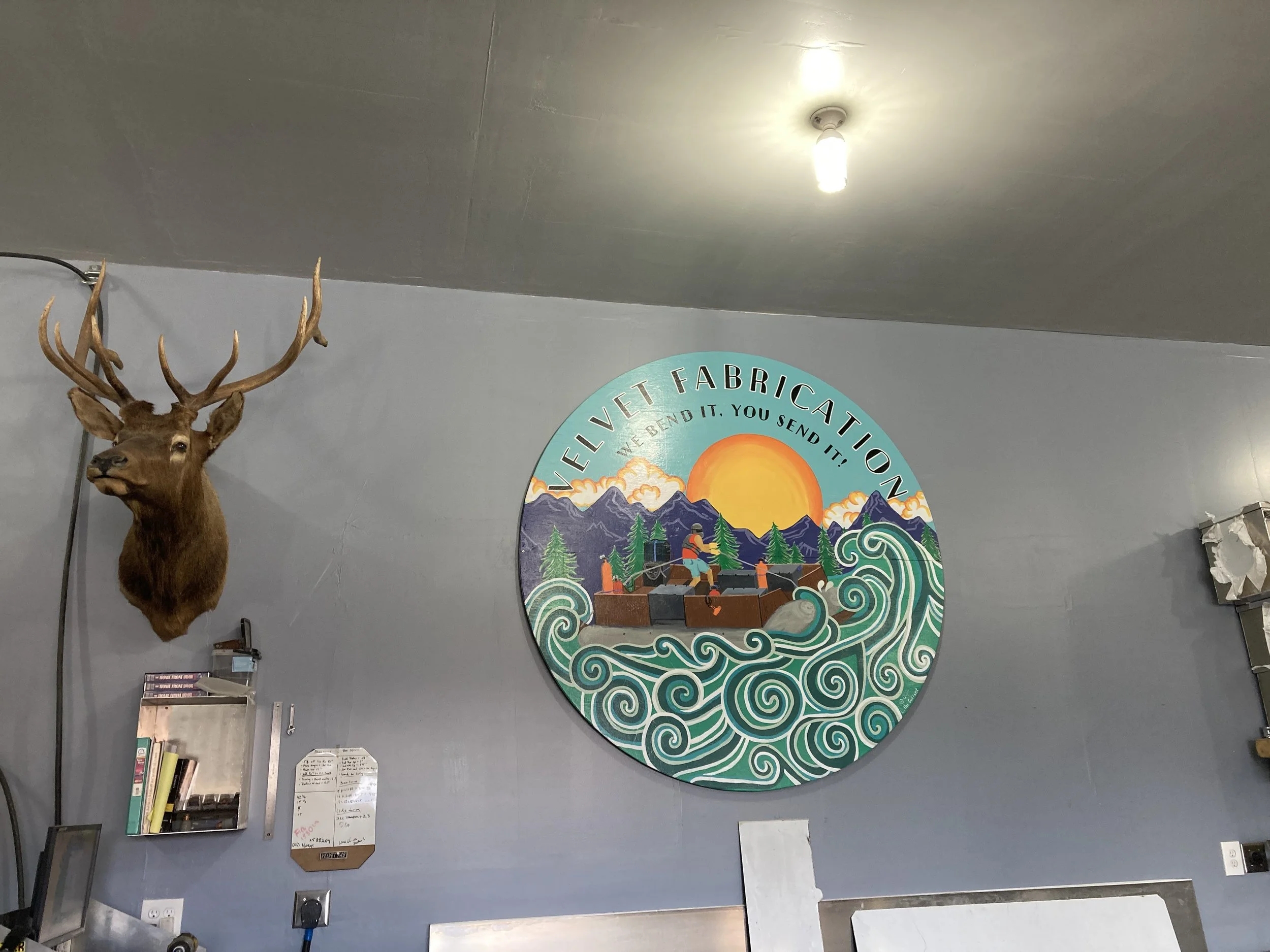 Interior of a building with a mounted deer head on the left and a colorful circular sign on the right. The sign features an illustration of mountains, pine trees, ocean waves, and a sunset, with the text 'Velvet Fabrication' and the slogan 'We Bend I