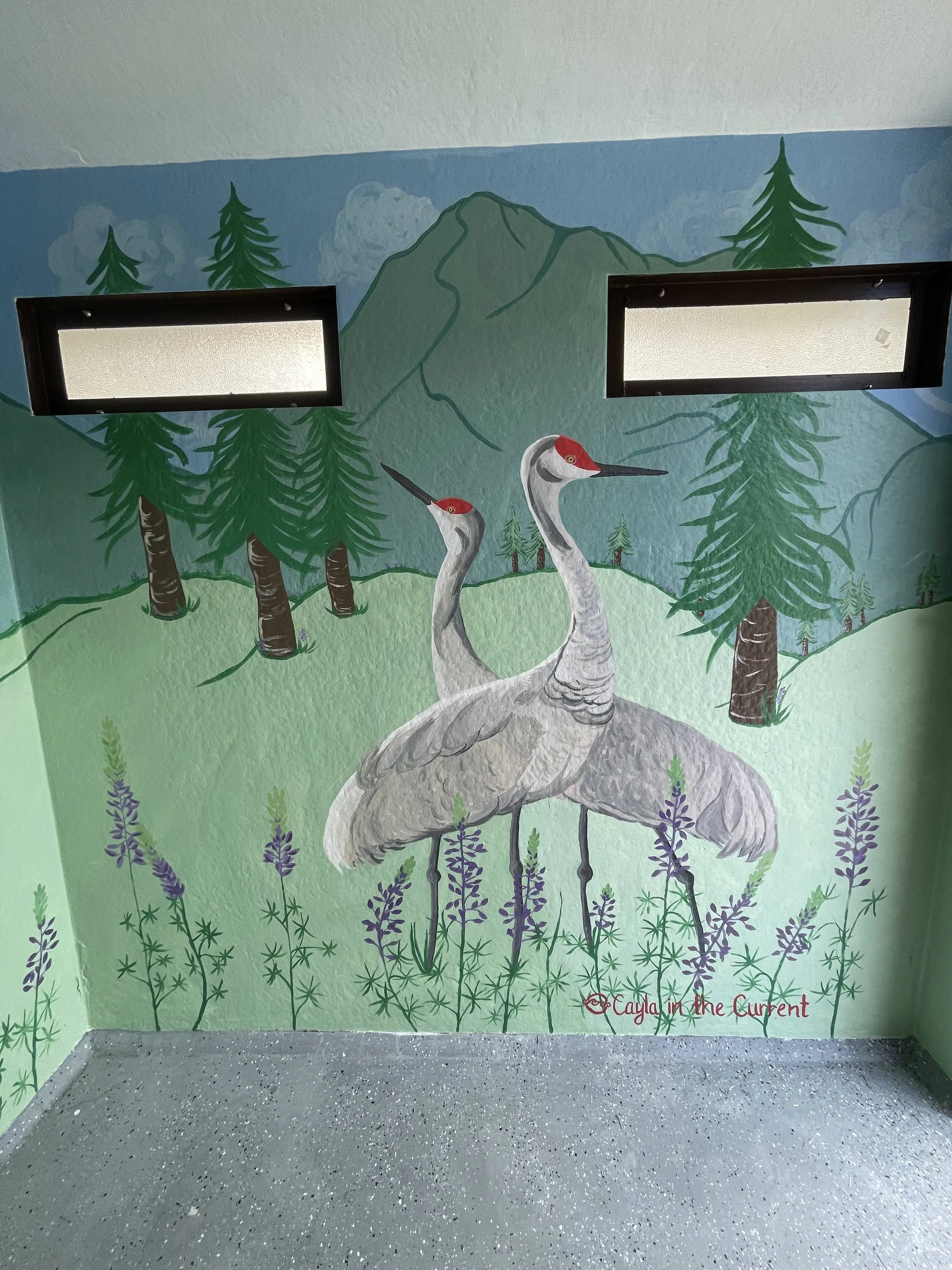 Mural painting of two cranes with red crowns standing among purple flowers in a forest landscape with green trees, mountain in the background, and two windows on the wall.