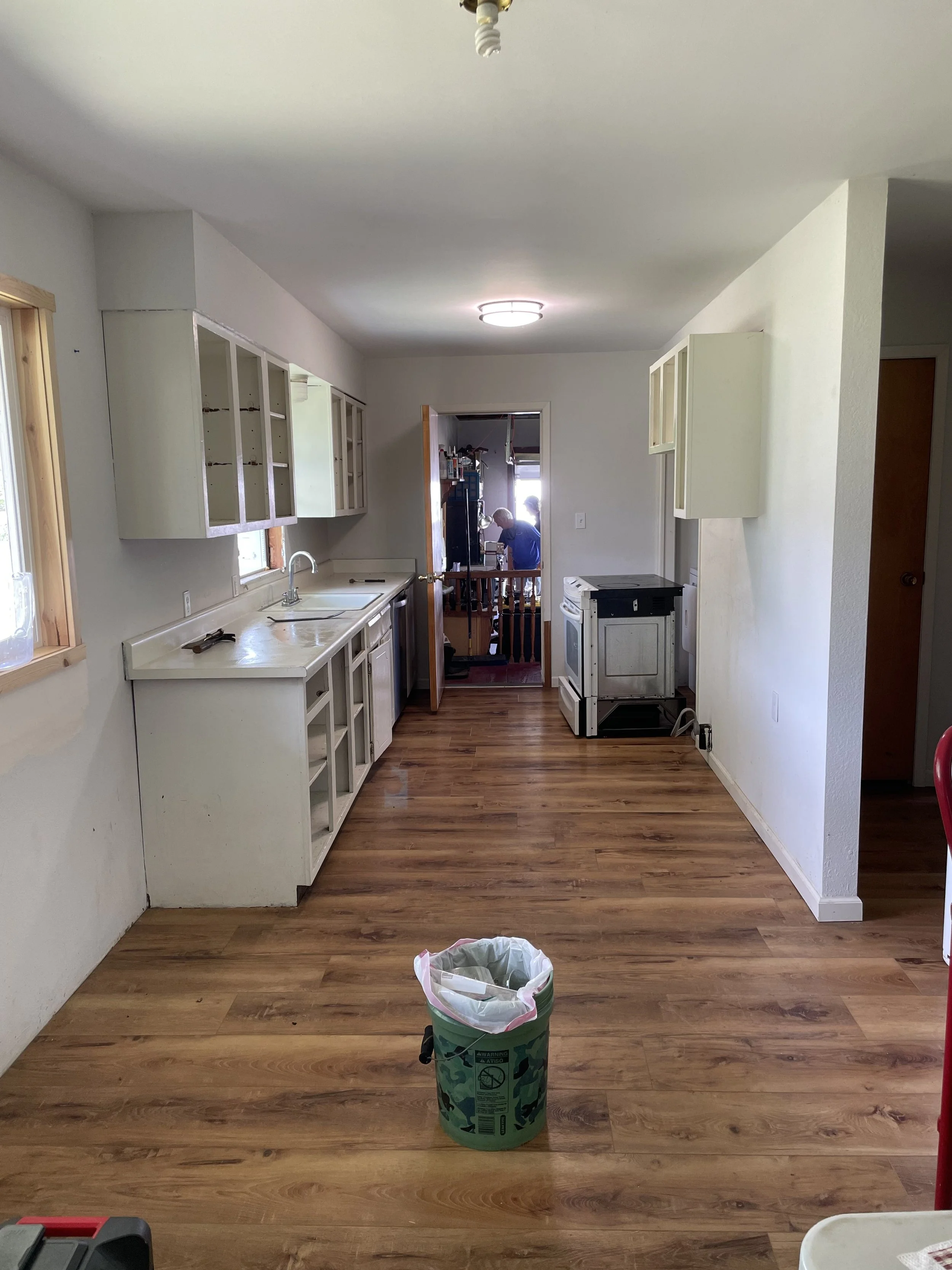 Empty kitchen with wooden floor, white cabinets, sink, and small window, with a trash can in the foreground and decorative light fixtures on the ceiling.