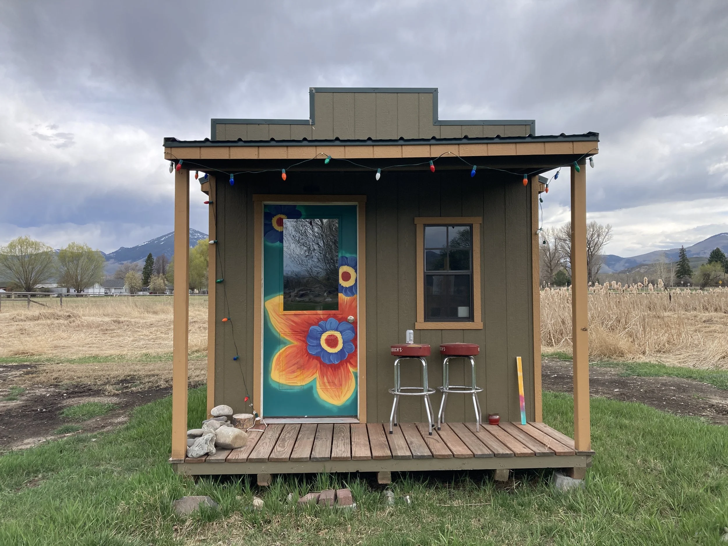 Small wooden porch with a painted door featuring a large orange and yellow butterfly with blue flowers, two red bar stools, and a colorful wooden stick, set against a rural landscape with mountains and cloudy sky.