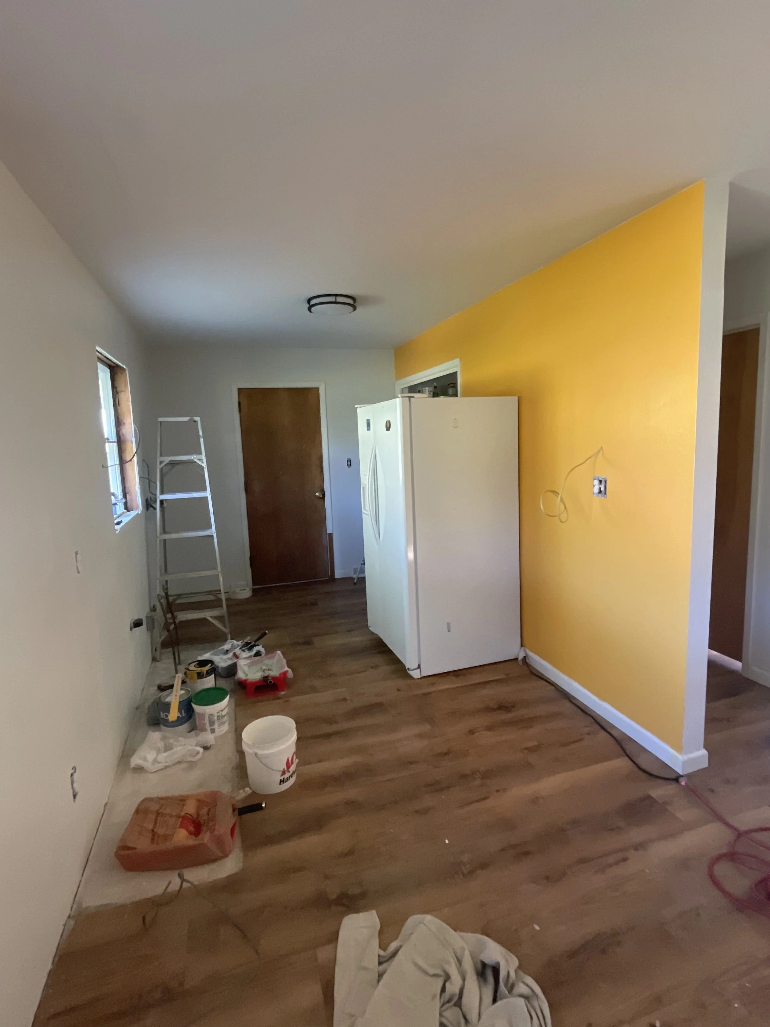 Living room under construction or renovation with tools and supplies on the floor, a yellow accent wall, a white refrigerator, a ladder, and a wooden door.