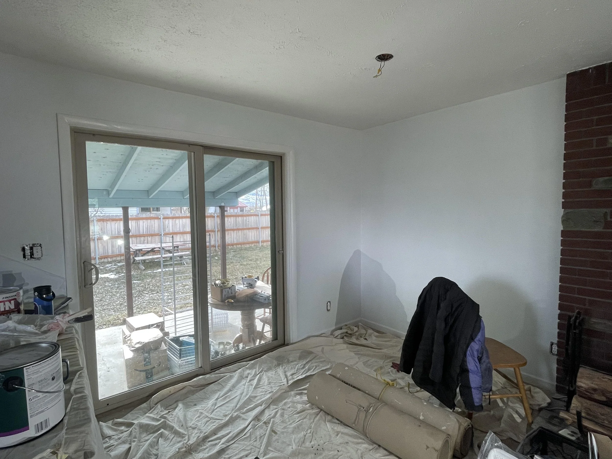 Room under renovation with a sliding glass door leading to an outdoor area, construction materials and tools scattered inside, a black jacket draped over a chair, and an unlit ceiling light fixture.