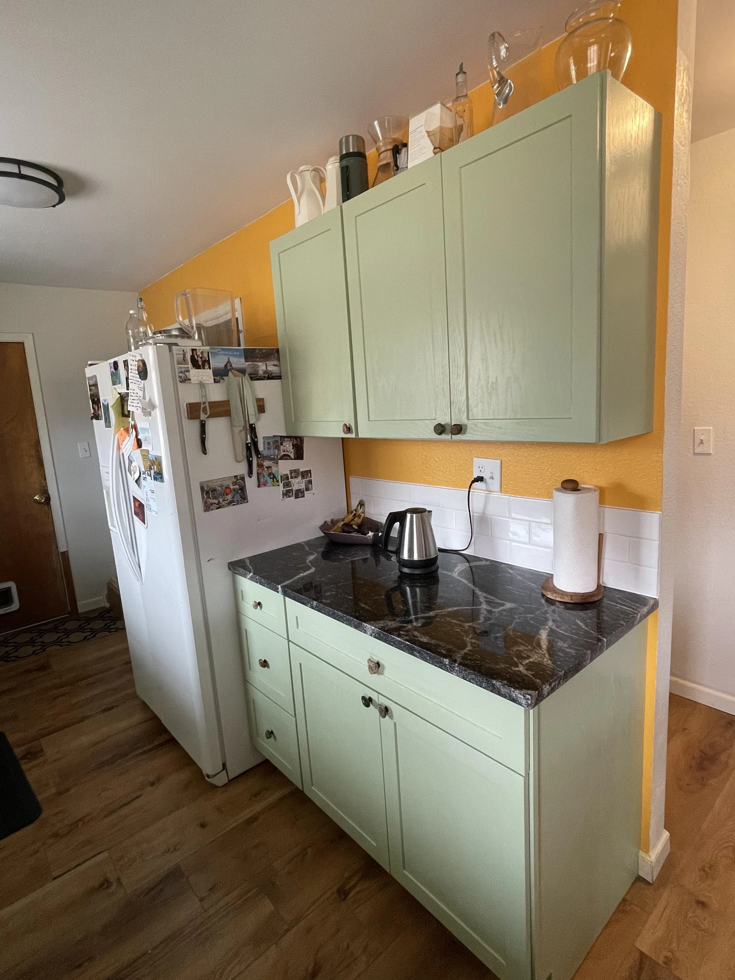 A kitchen corner with light green cabinets, a black marble countertop, a white refrigerator decorated with photos and magnets, and a yellow wall with various kitchen items on top of the cabinets.