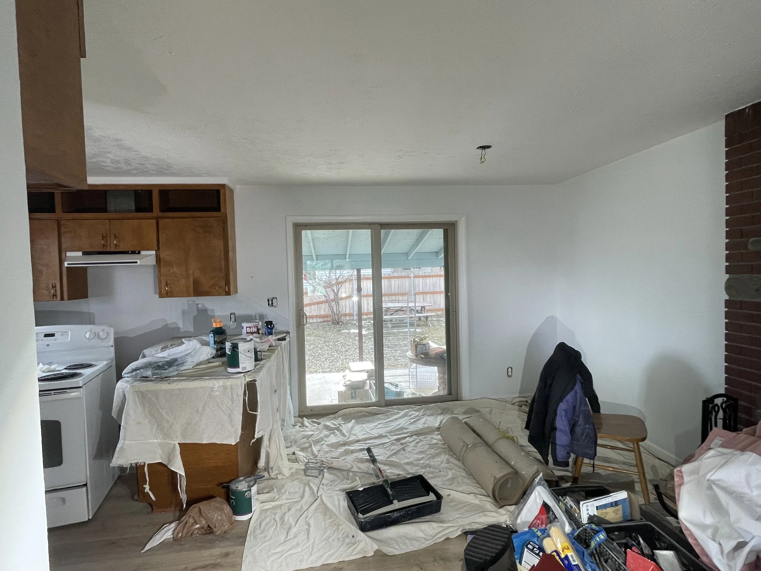 Living room under renovation with tools, paint cans, and supplies scattered on a covered surface, a sliding glass door leading outside, and furniture covered with drop cloths.