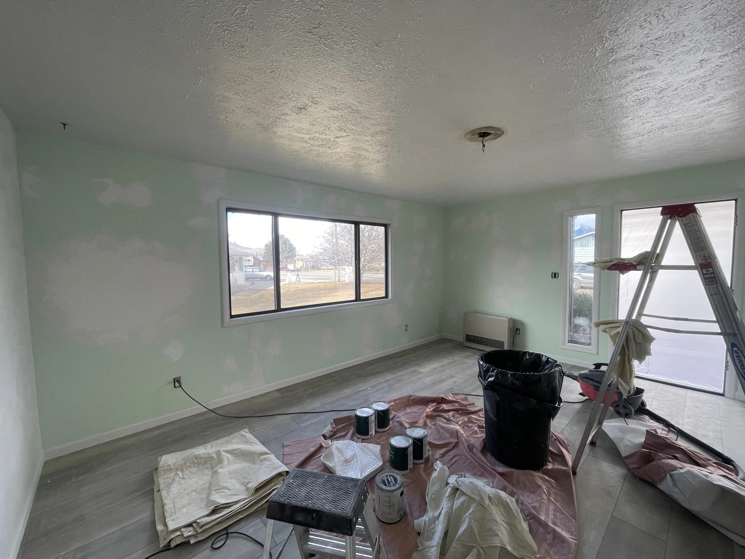 Living room under renovation with light green painted walls, large window, ladder, and painting supplies on the floor.