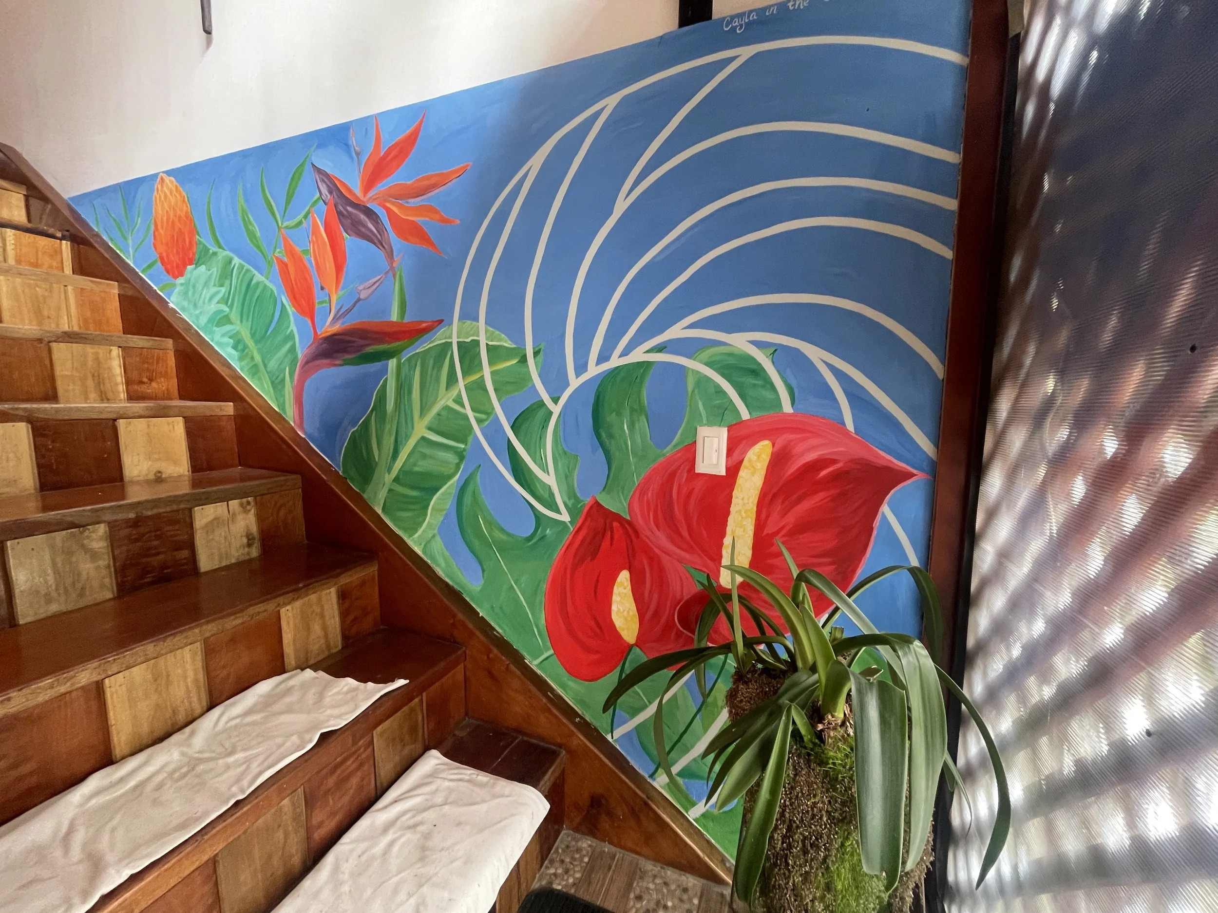 A colorful mural on a staircase wall featuring tropical flowers and large green leaves, with a potted plant placed in front of it.