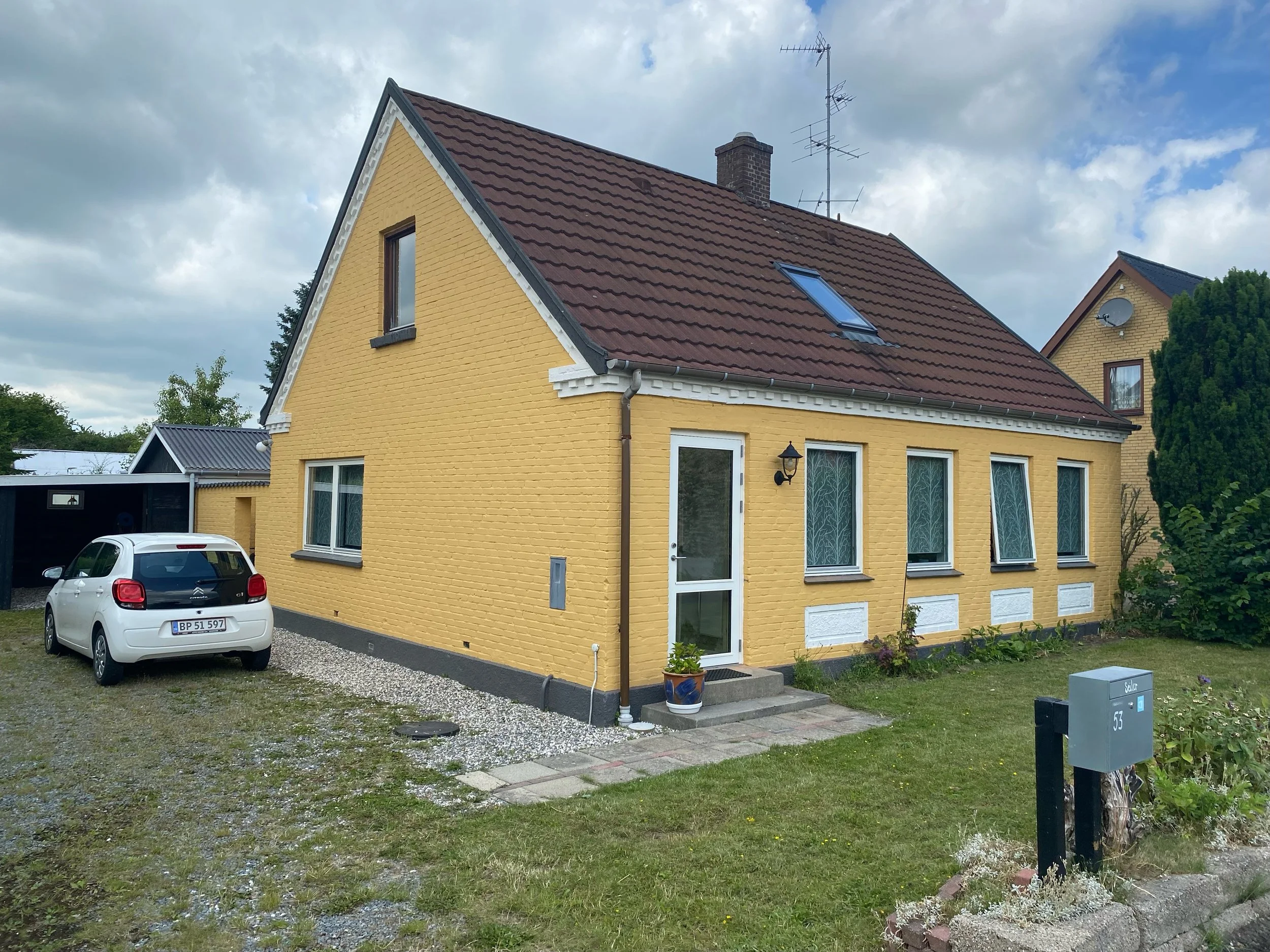 A yellow house with a red-tiled roof, four front windows, a front door with a small step, and a potted plant outside. A white car is parked on a gravel driveway beside the house. The lawn surrounds the house, and there is a mailbox with the house num