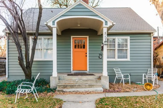 A house with a light blue exterior, a front porch with stairs, an orange front door, and two windows with white shutters. There are two white chairs on the right side of the porch and a white chair on the left side in the yard. A tree is on the left 