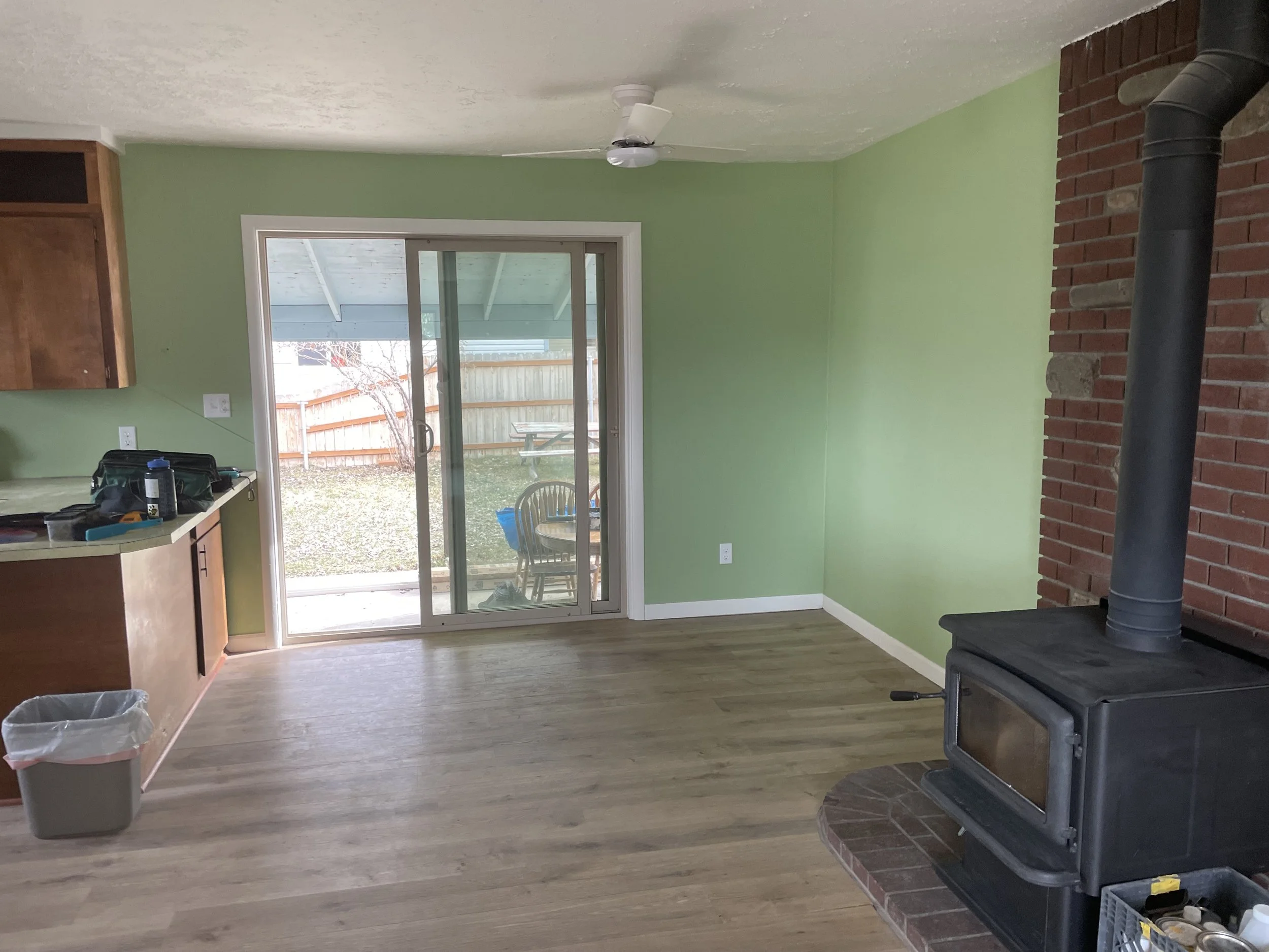 Empty living room with green walls, hardwood floors, sliding glass door leading to backyard, brick fireplace with stove, ceiling fan, and some clutter on the kitchen counter.