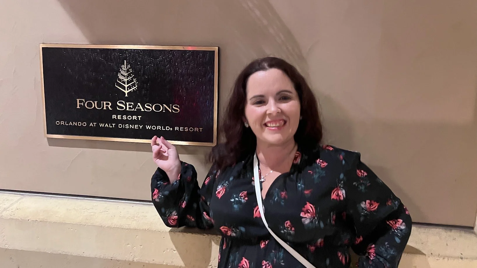 A woman with shoulder-length dark hair, wearing a black floral dress, smiling while pointing at a sign for the Four Seasons Resort at Walt Disney World in Orlando.