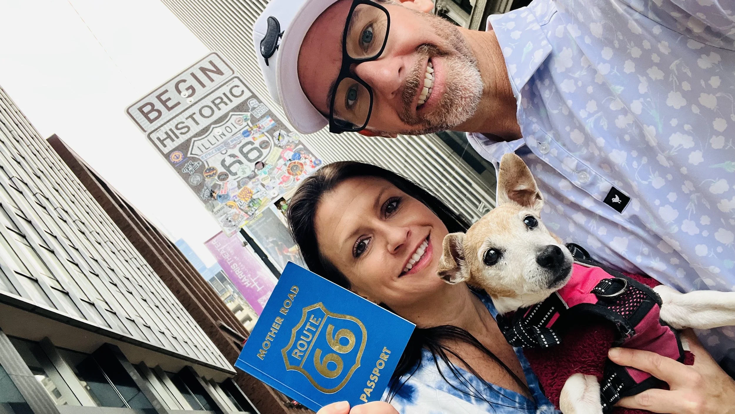 A smiling man with glasses and a white hat, a smiling woman holding a small dog, and a man holding a blue sign that says 'Route 66 Mother Road' in front of brick buildings.
