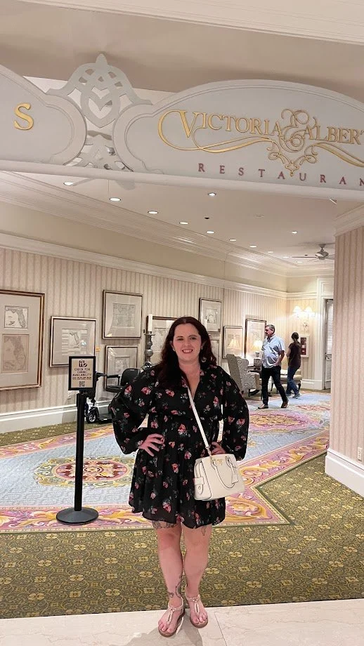 Woman in floral dress with white purse standing inside Victoria & Albert restaurant, with framed artwork on walls and other patrons in the background.