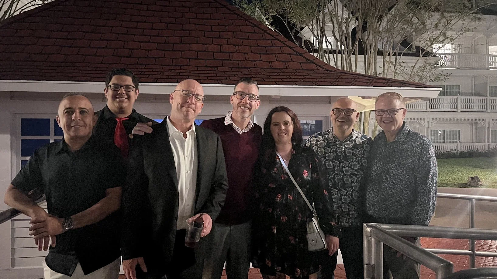 Group of seven people standing outside at night in front of a building with trees. They are dressed in a mix of formal and casual attire, smiling at the camera.