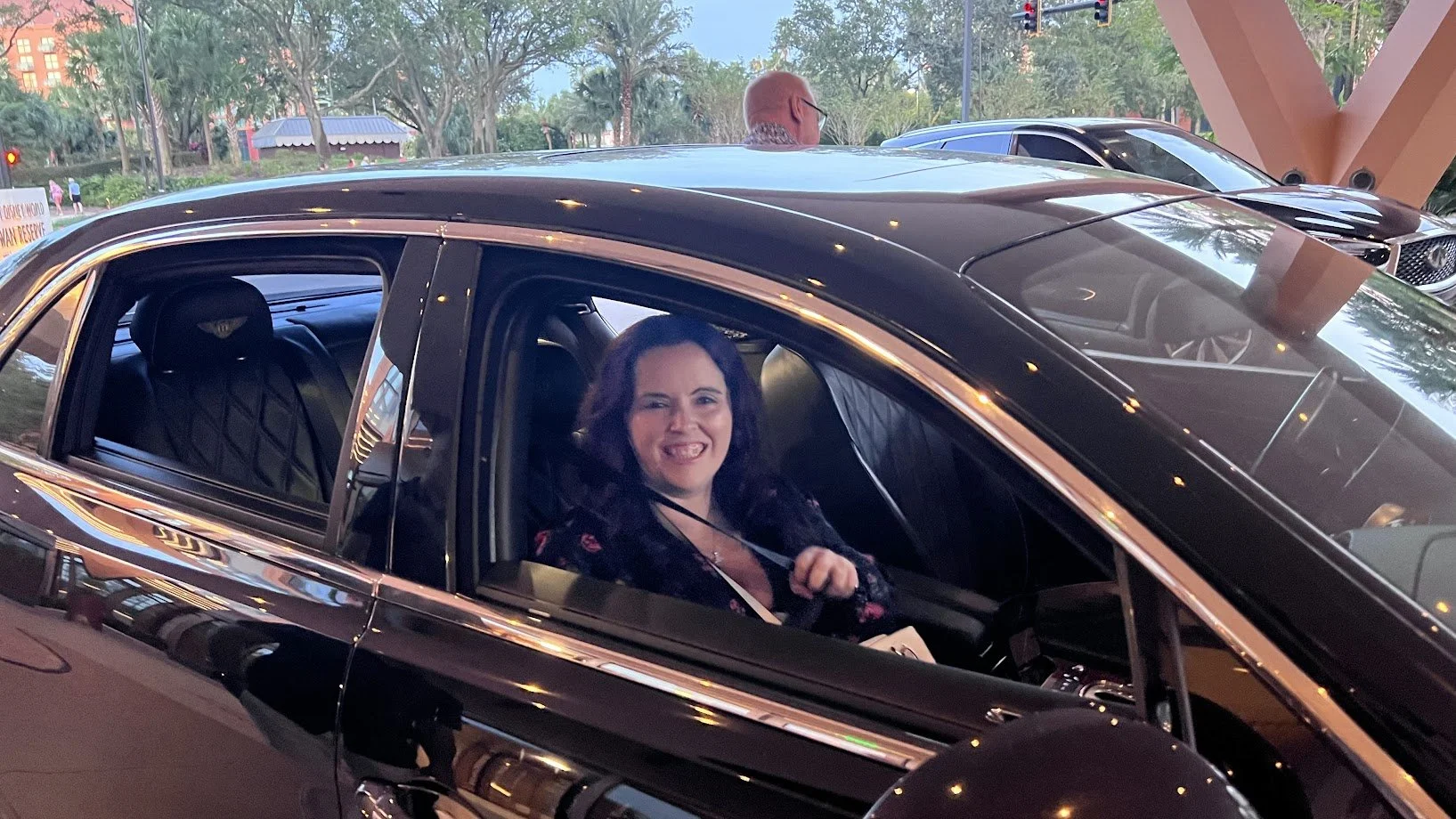 Woman sitting in a black luxury car, smiling and looking through the open car window, with other cars and trees in the background.