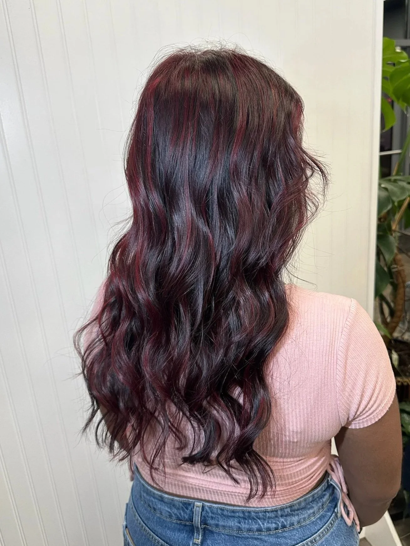 Back view of a woman with long, wavy hair that has dark base color with red highlights, wearing a light pink shirt and blue jeans.