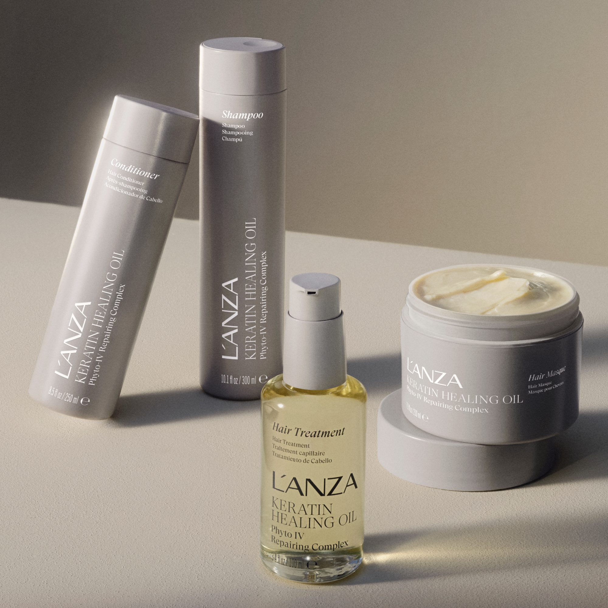 A collection of hair care products including a jar of L'ANZA Keratin Healing Oil hair mask, a bottle of L'ANZA Keratin Healing Oil spray, a bottle of L'ANZA Keratin Healing Oil conditioner, and a bottle of L'ANZA Keratin Healing Oil shampoo placed on a light surface with a neutral background.