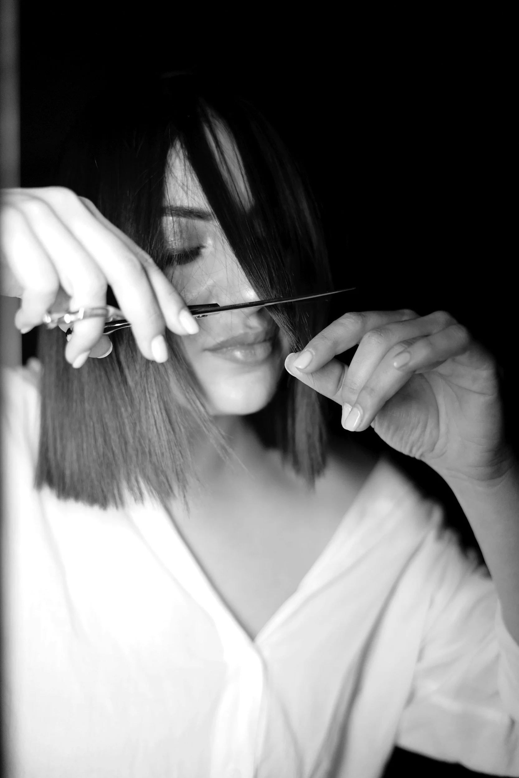 Black and white photo of a woman with short dark hair, holding a pair of scissors near her face, with one eye closed, in a dark background.