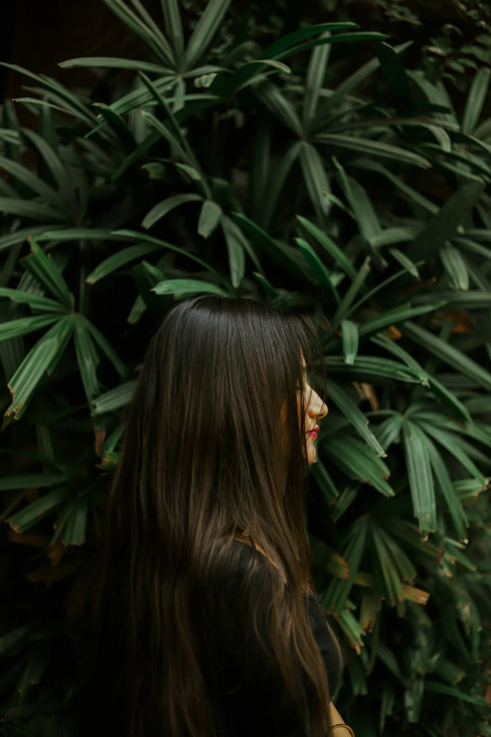 A woman with long dark hair and pink lipstick standing in front of lush green tropical plants.