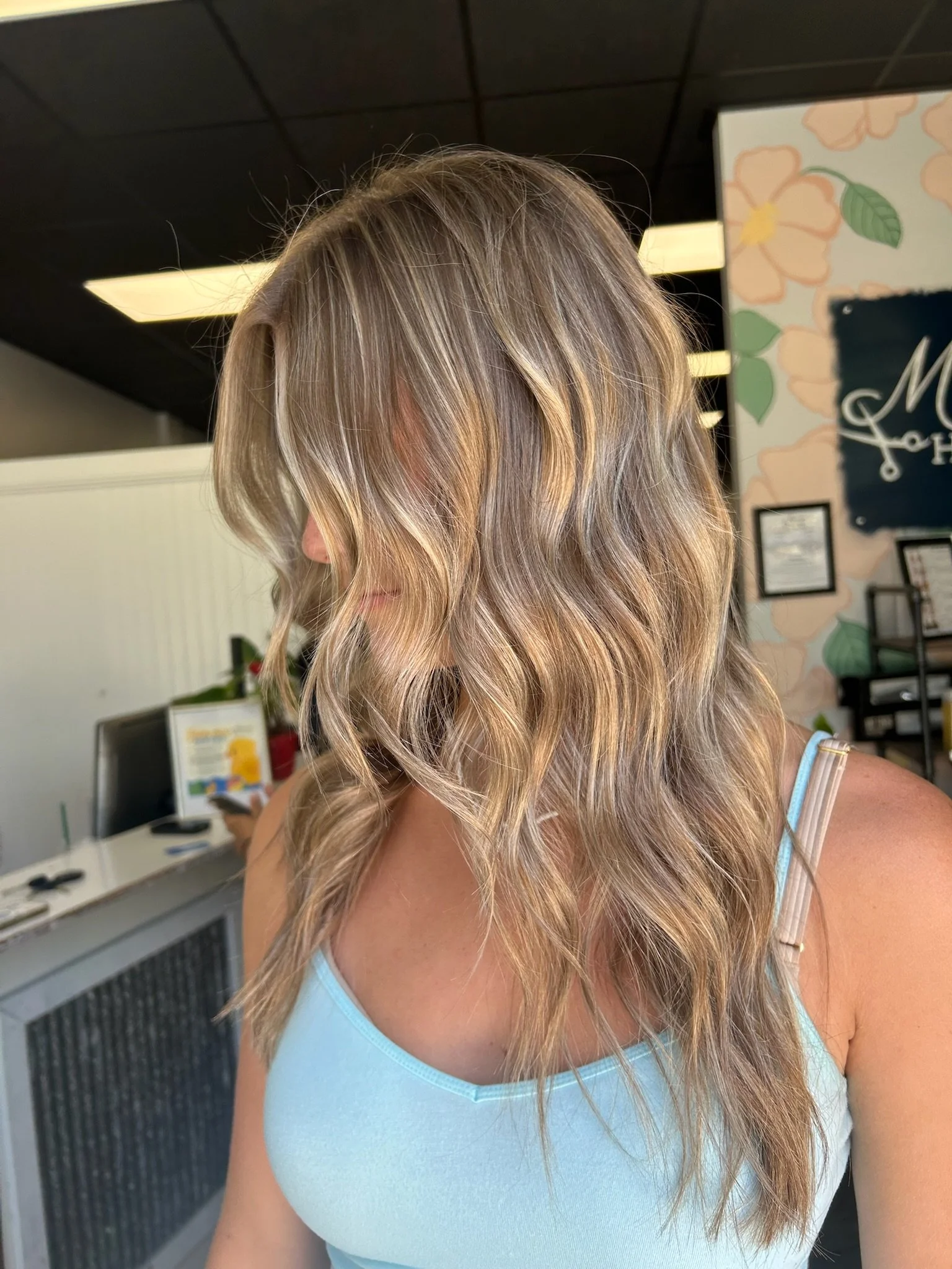 A woman with wavy blonde hair in a salon with a floral mural, reception desk, and a person in the background.