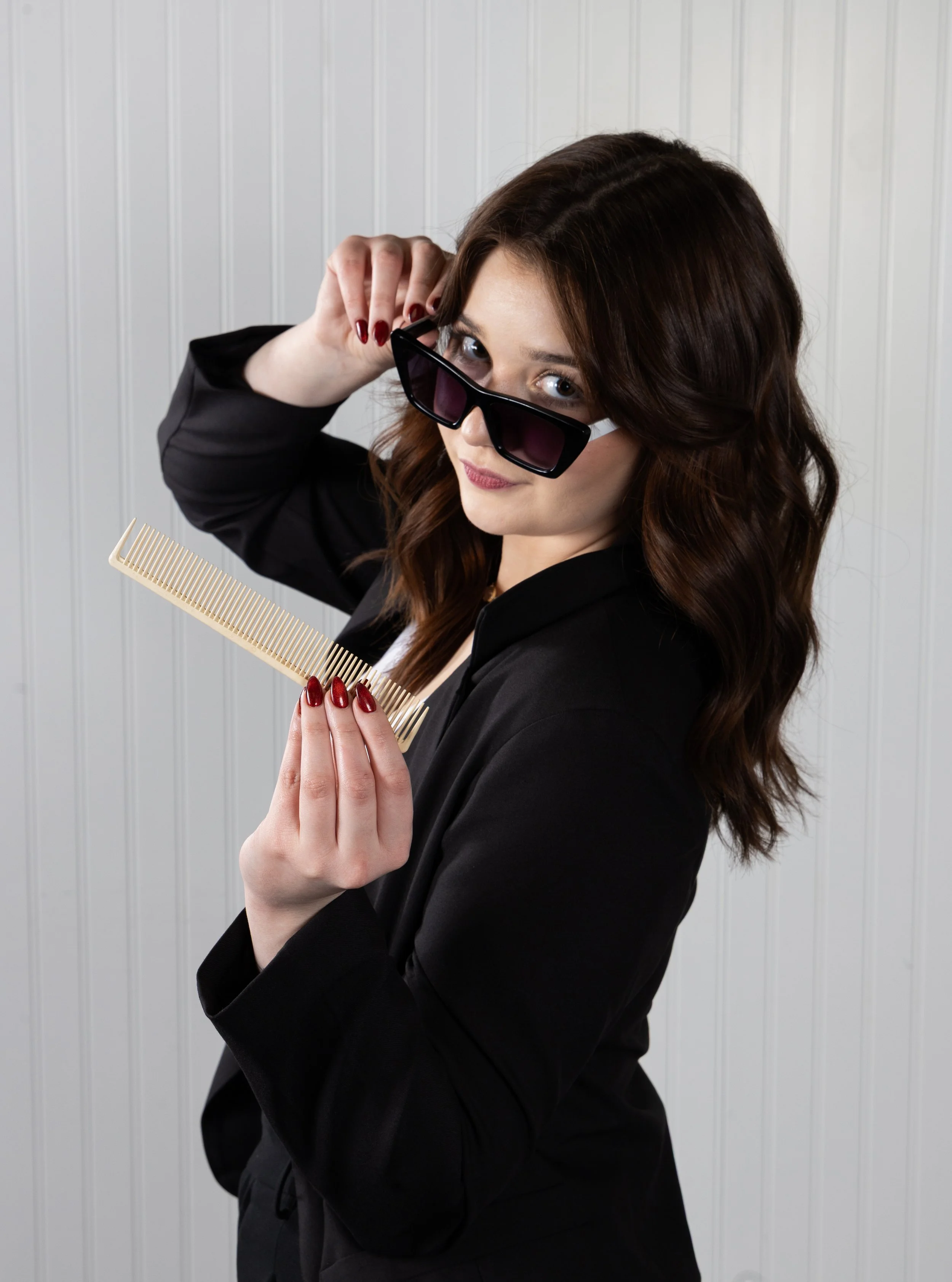 A woman with wavy brown hair, wearing a black blazer and red lipstick, holding a wide tooth comb and adjusting her sunglasses while looking at the camera against a white paneled background.