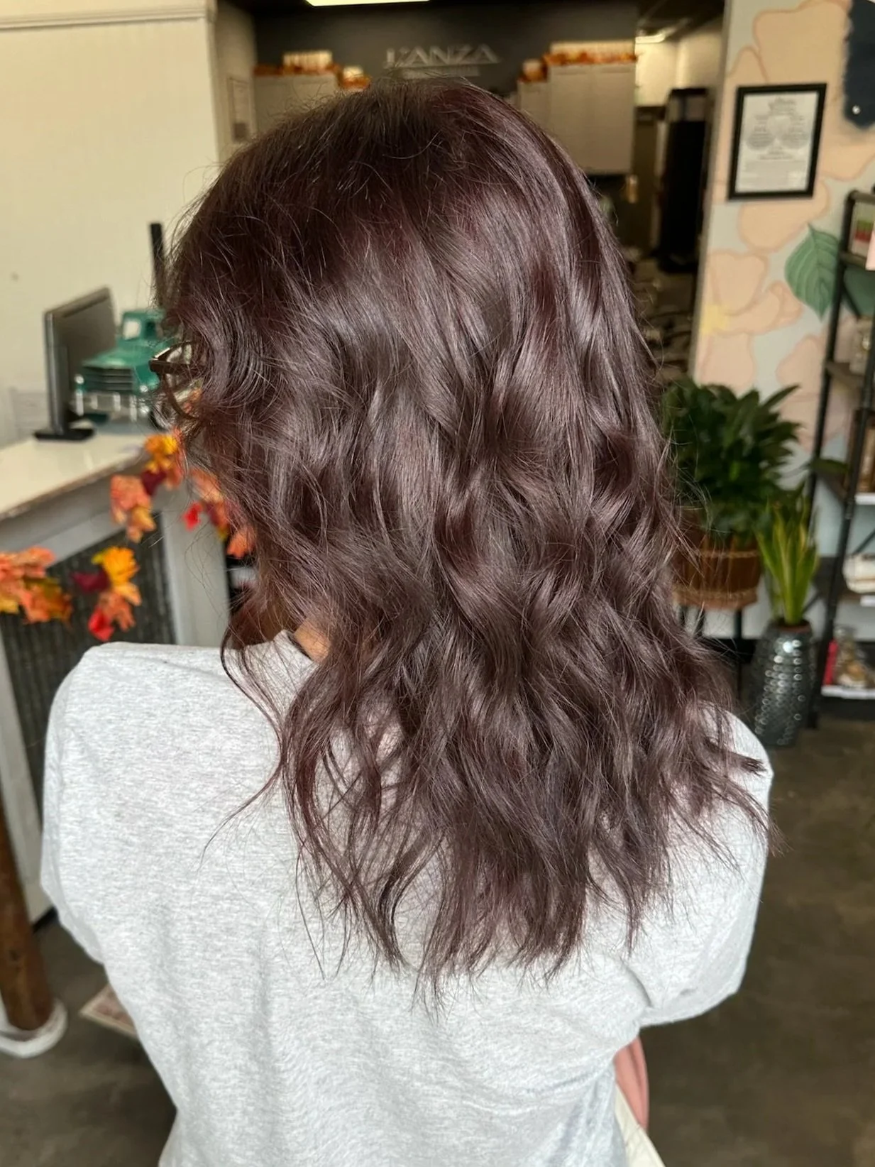 Back view of a person with shoulder-length wavy brunette hair inside a salon.