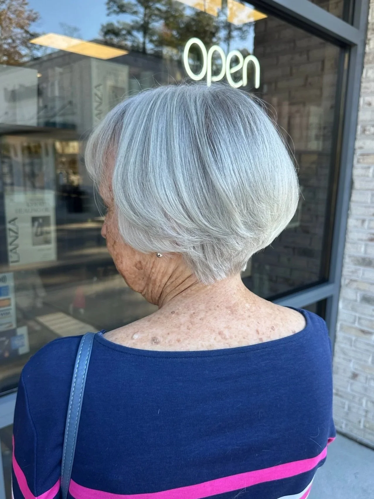 Older woman with short gray hair looking at a store window with a neon 'Open' sign, wearing a navy blue top with pink stripes.