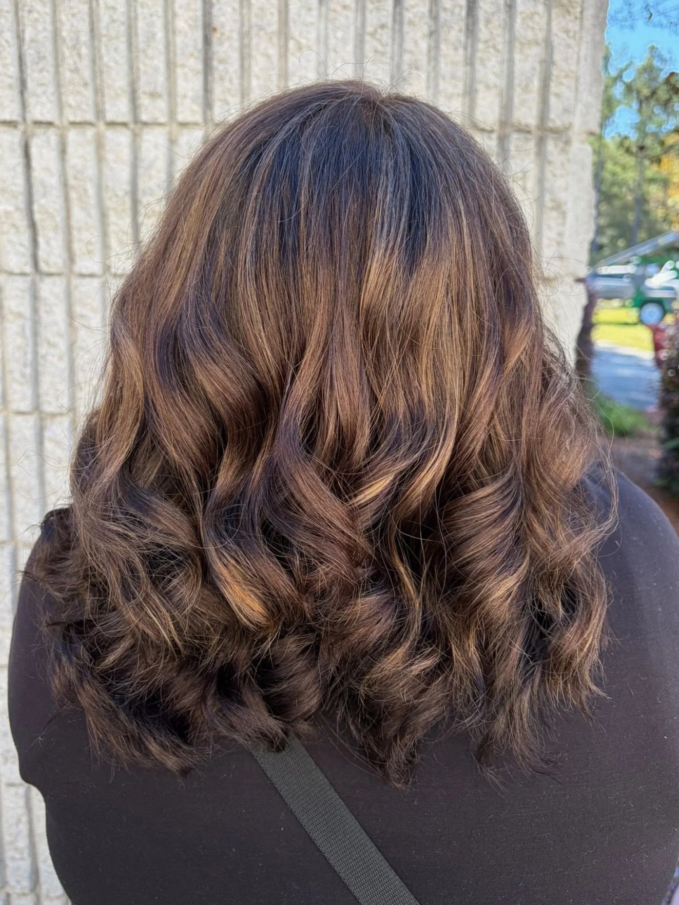 Back view of a woman with shoulder-length wavy hair featuring brown and blonde highlights, standing outdoors near a beige brick wall.