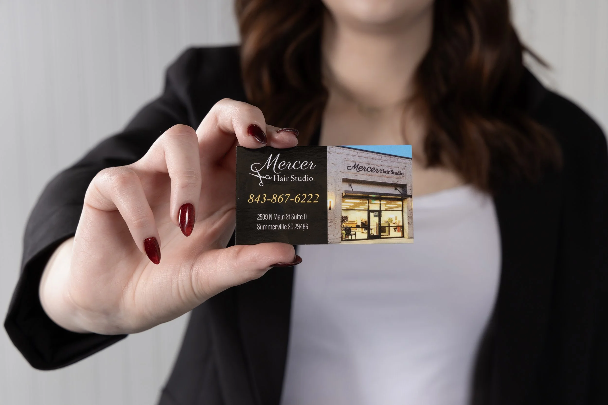 A woman with red nail polish holding a business card for Mercer Hair Studio, which includes a photo of the salon, phone number 843-867-6222, and address 2509 N Main St Suite D, Summerville SC 29486.