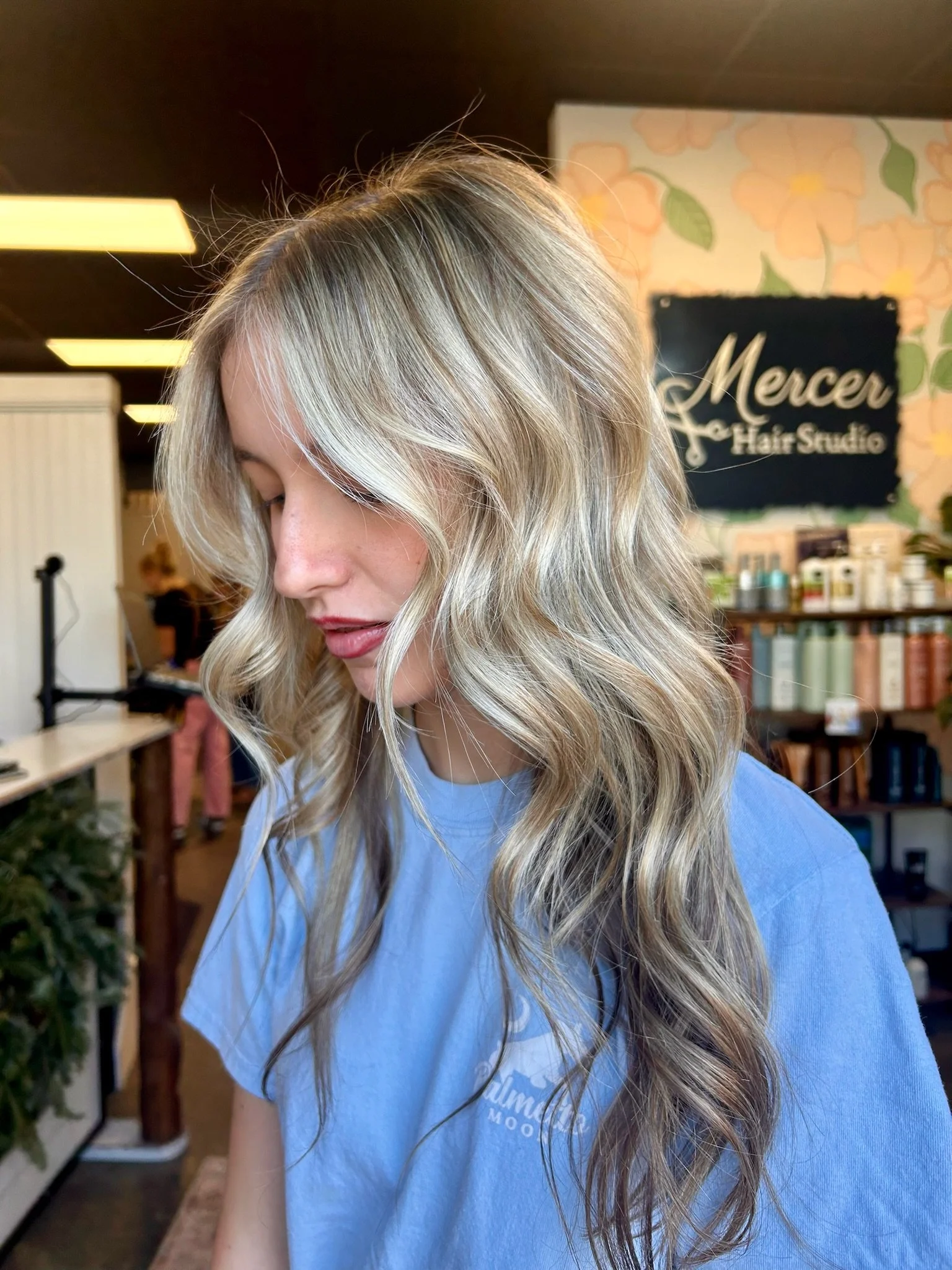 A woman with long, wavy blonde hair, wearing a light blue T-shirt, standing in a hair salon.