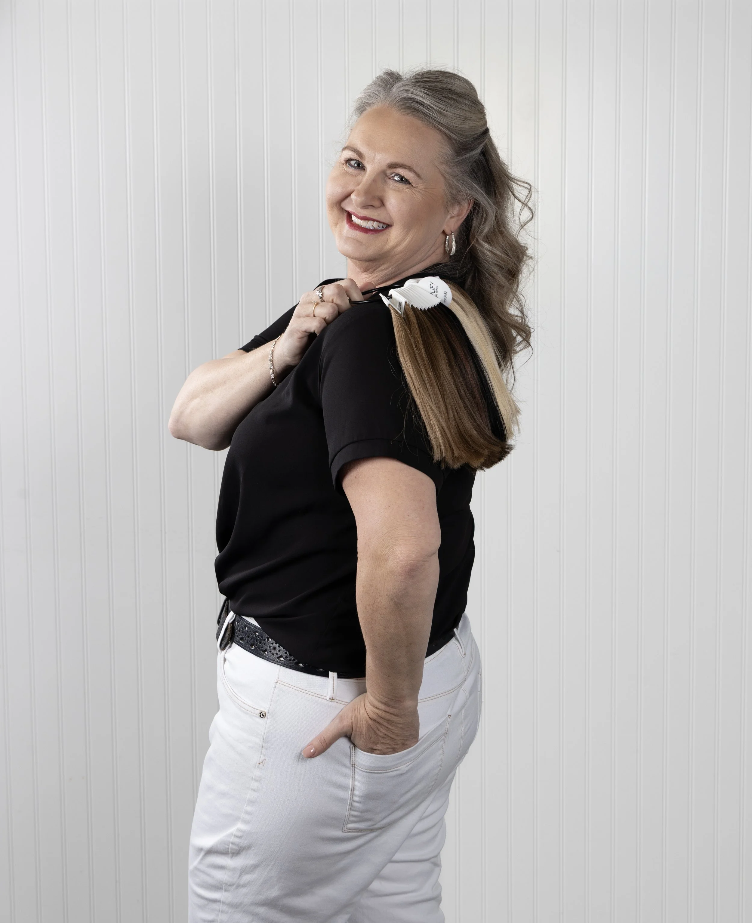 A smiling woman with gray hair, wearing a black shirt and white pants, holding a color swatch sample against her shoulder, standing in front of a white paneled wall.