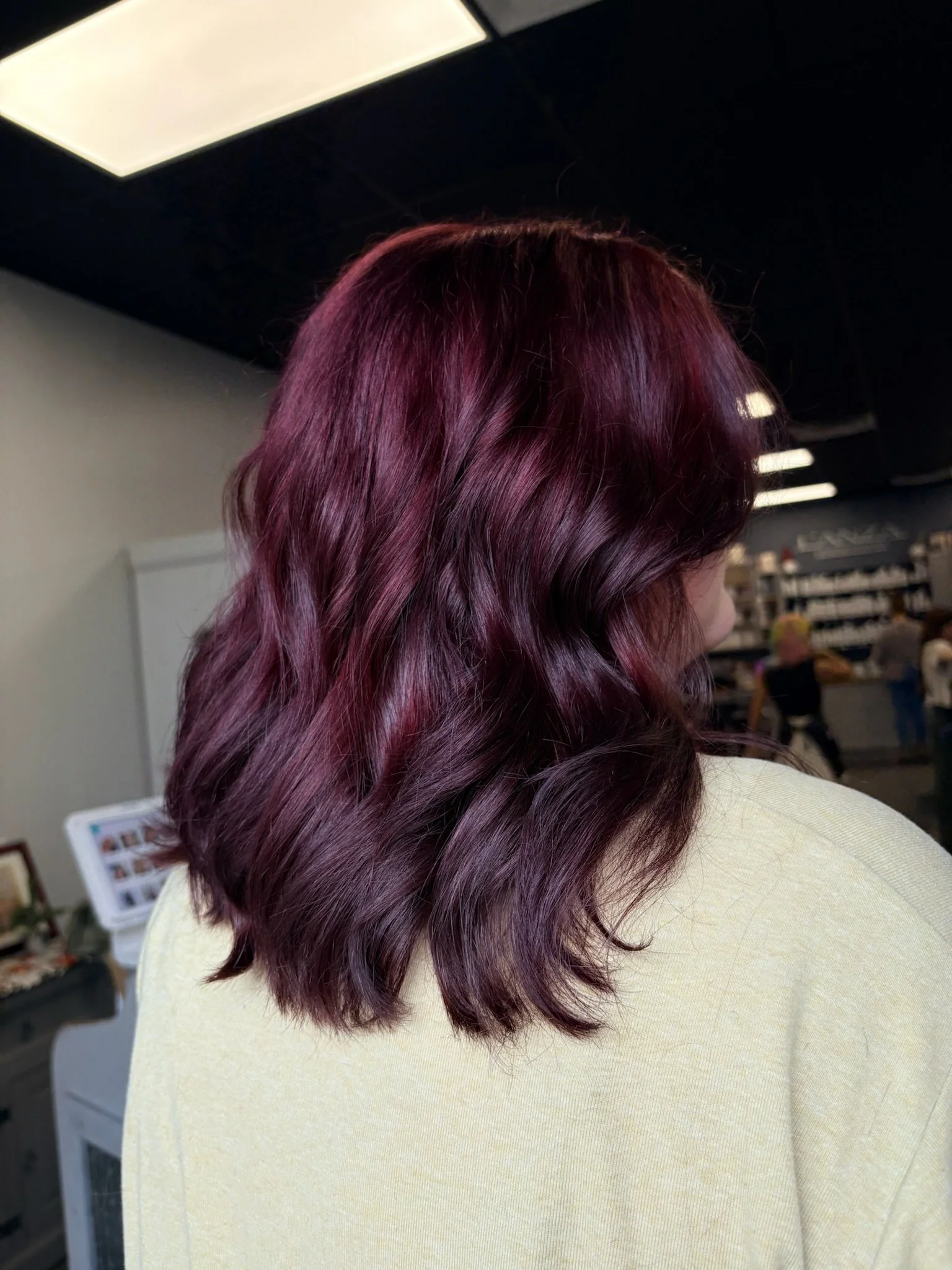 Woman with wavy, shoulder-length burgundy hair wearing a light yellow top, indoors at a salon or store.