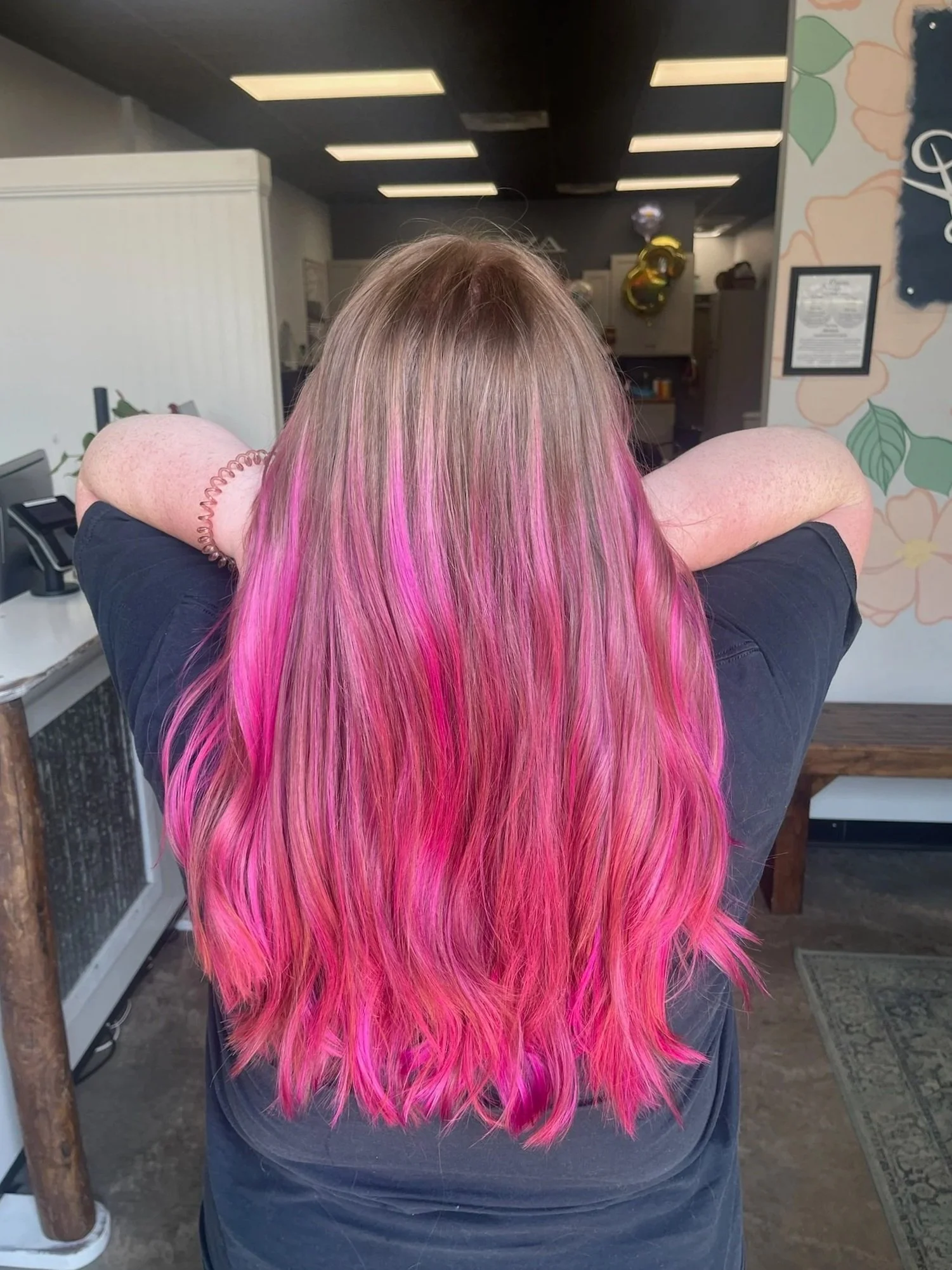 Person with long, wavy hair dyed in shades of pink, sitting with arms crossed behind their head inside a salon or studio.
