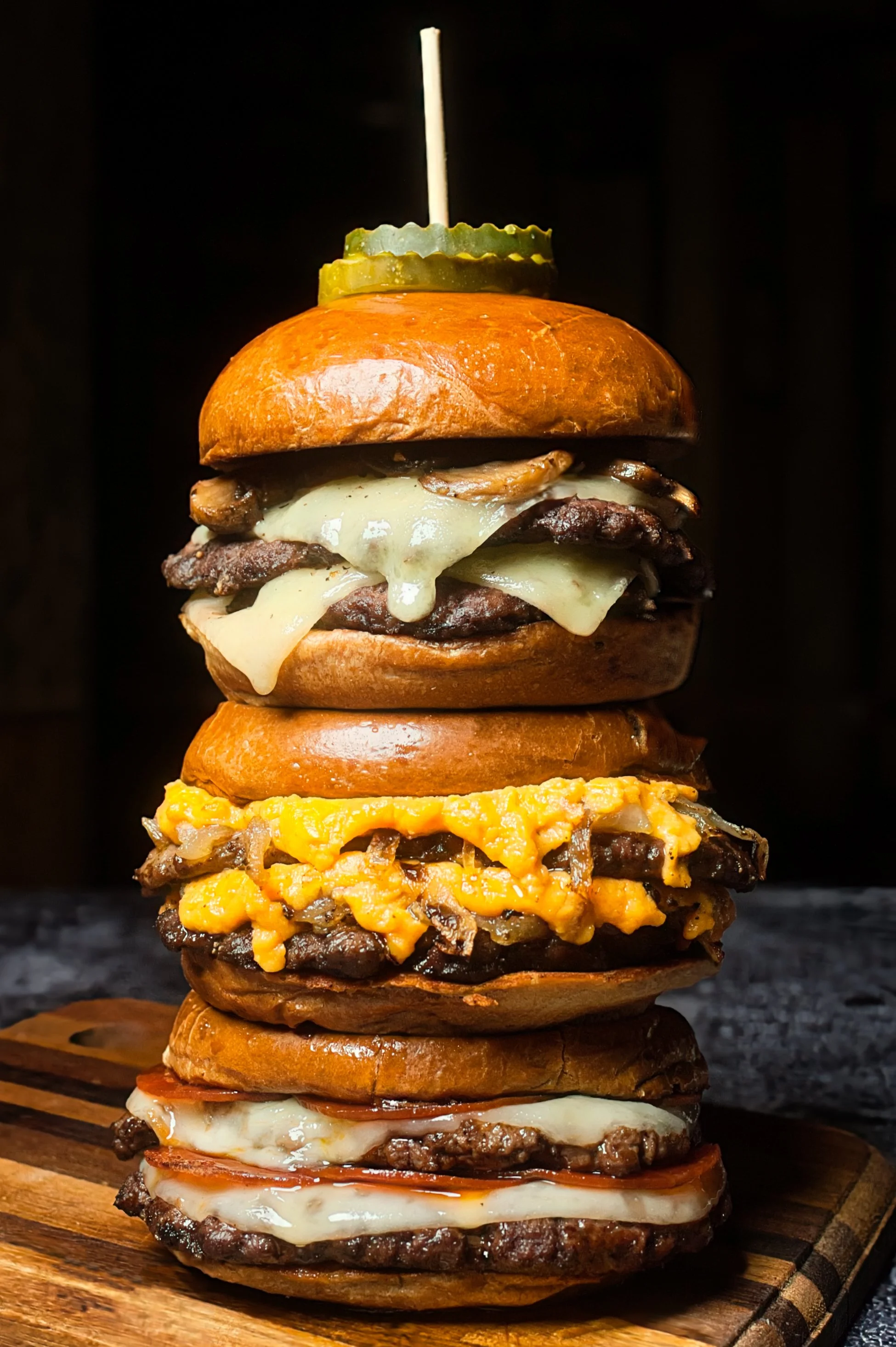Stacked gourmet burgers with various toppings including cheese, hot honey pepperoni, mushrooms, pickles, and sauces on a wooden board against a dark background.