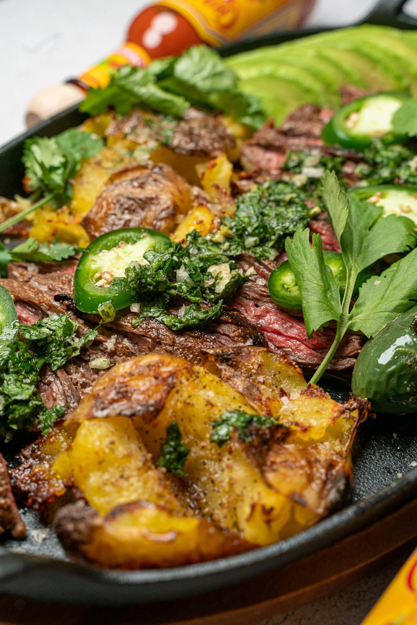 Close-up of a cast iron skillet filled with seasoned grilled steak, roasted potatoes, fresh herbs, and sliced green jalapenos.