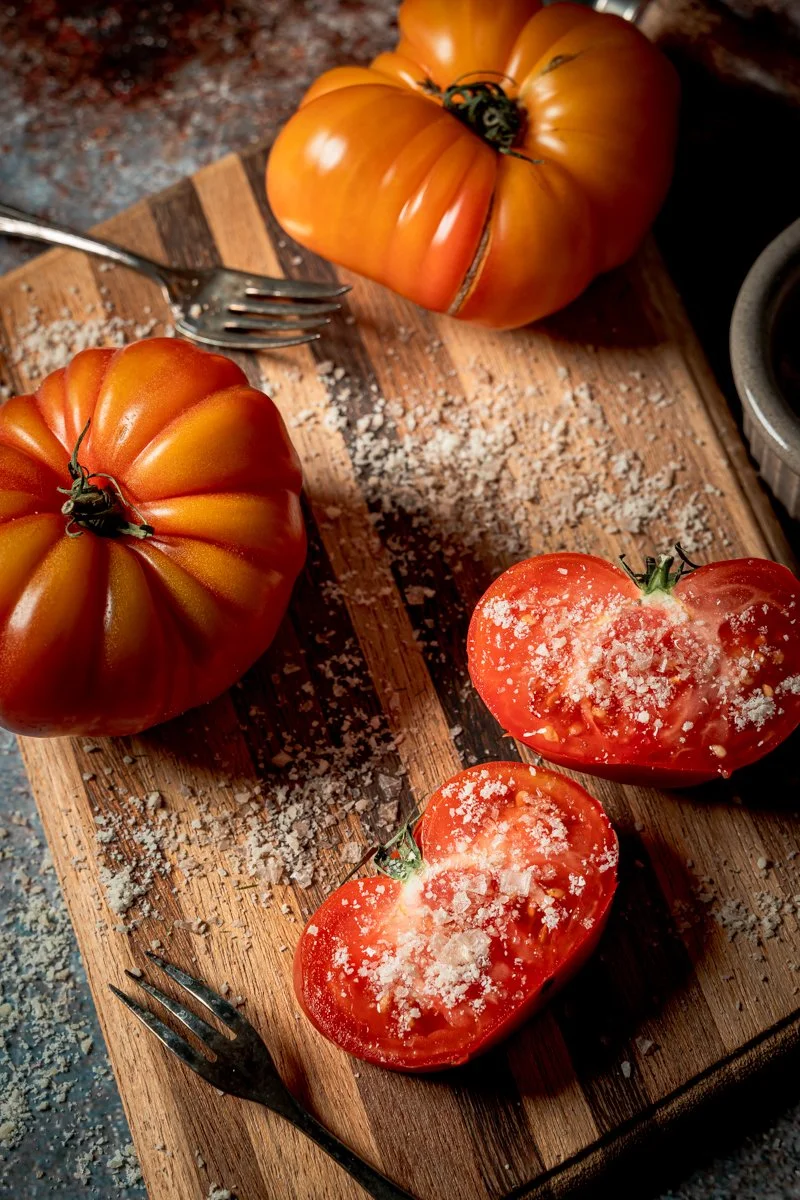 Two whole heirloom tomatoes and one sliced tomato topped with salt on a wooden cutting board, with a fork and parmesan scattered around.