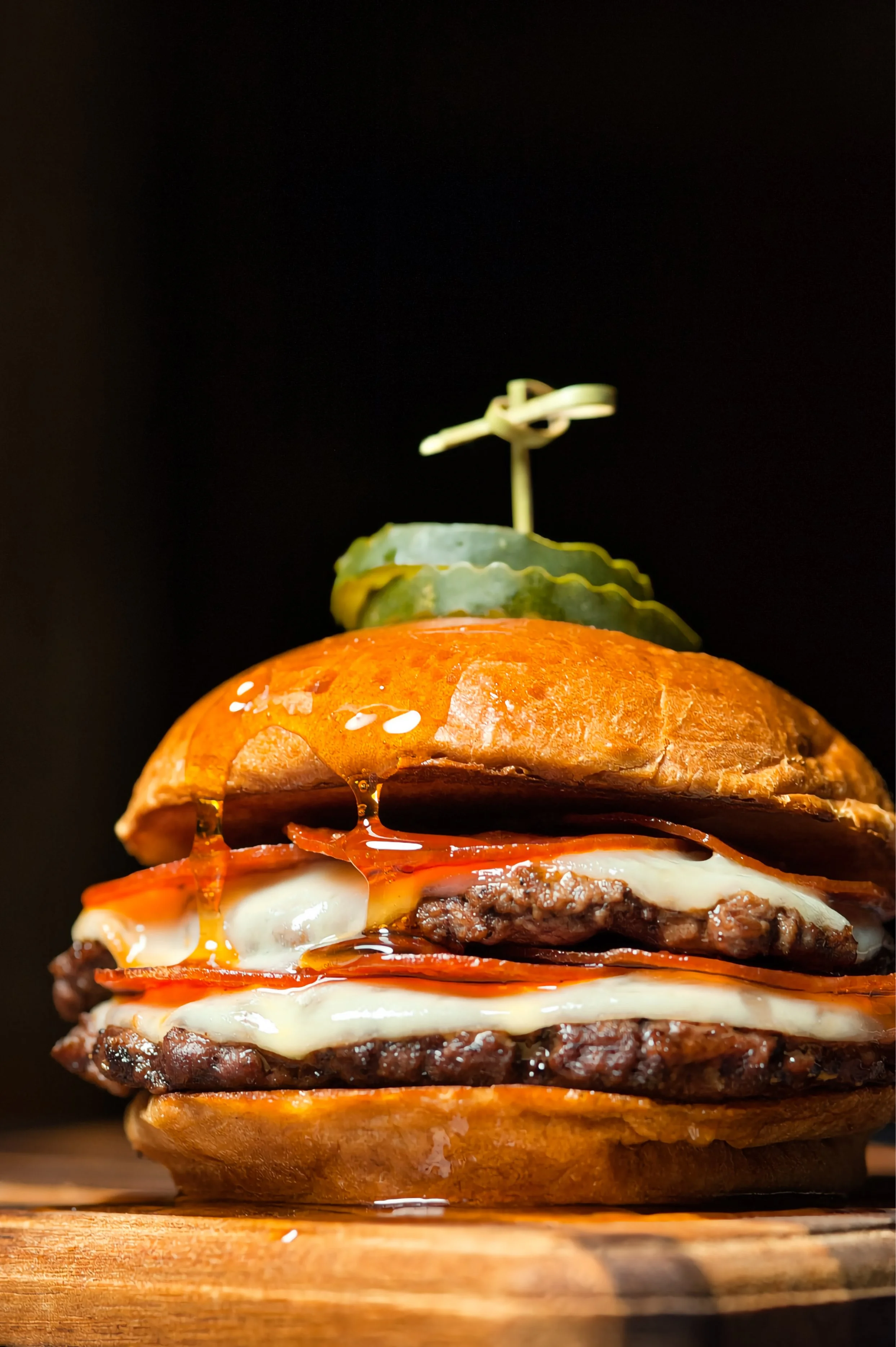 Close-up of a double  smashburger with melted cheese, lettuce, tomato, and hot honey pepperoni, topped with a pickle spear and toothpick against a black background.