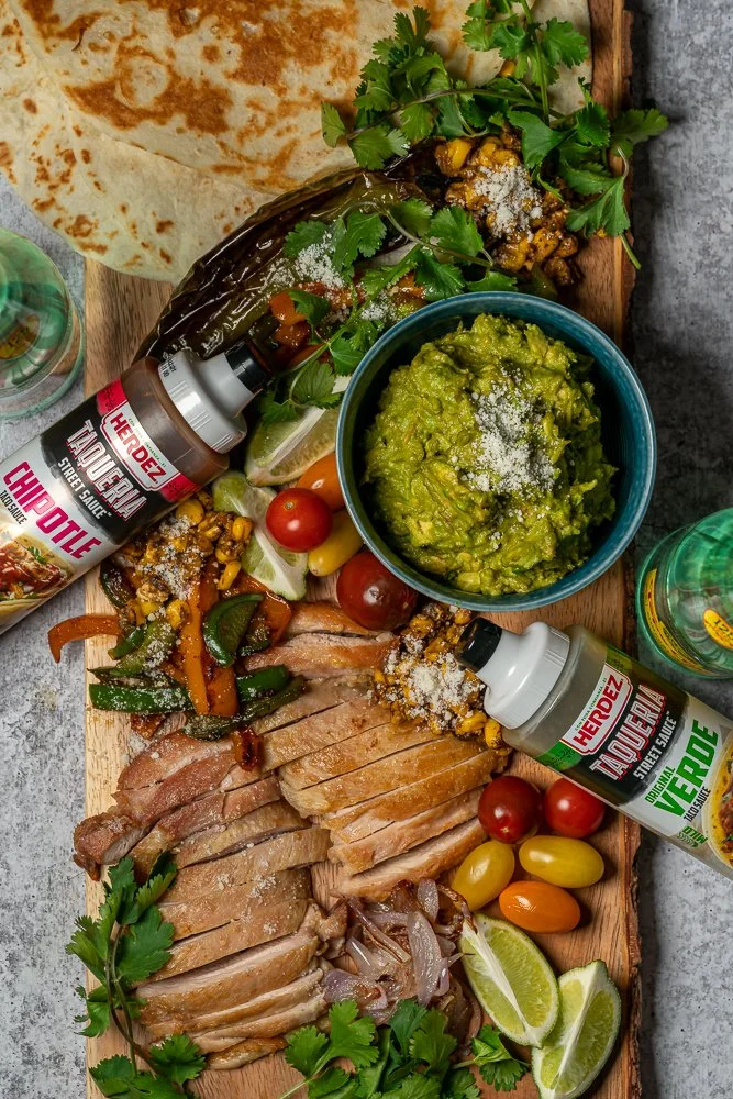 A wooden board with sliced grilled chicken, cherry and grape tomatoes, lime wedges, and cilantro, alongside a bowl of guacamole, grilled jalapeño pepper, grilled vegetables, and various sauces and beverages.