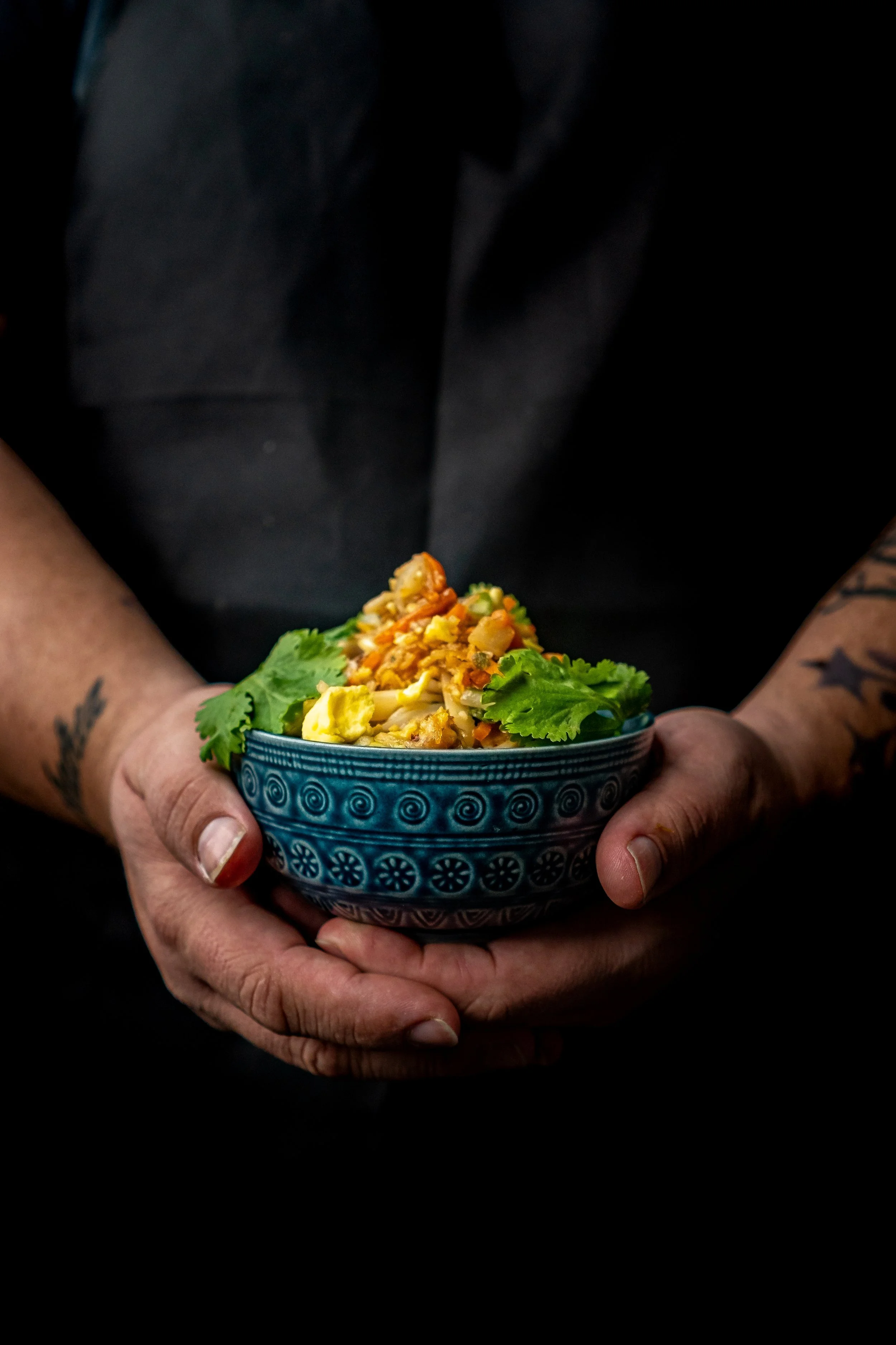 Vibrant Pad Thai garnished with fresh cilantro in a blue ceramic bowl, held by a chef in a dark apron. Dark and moody commercial food photography by Chicago Culinary Photo.