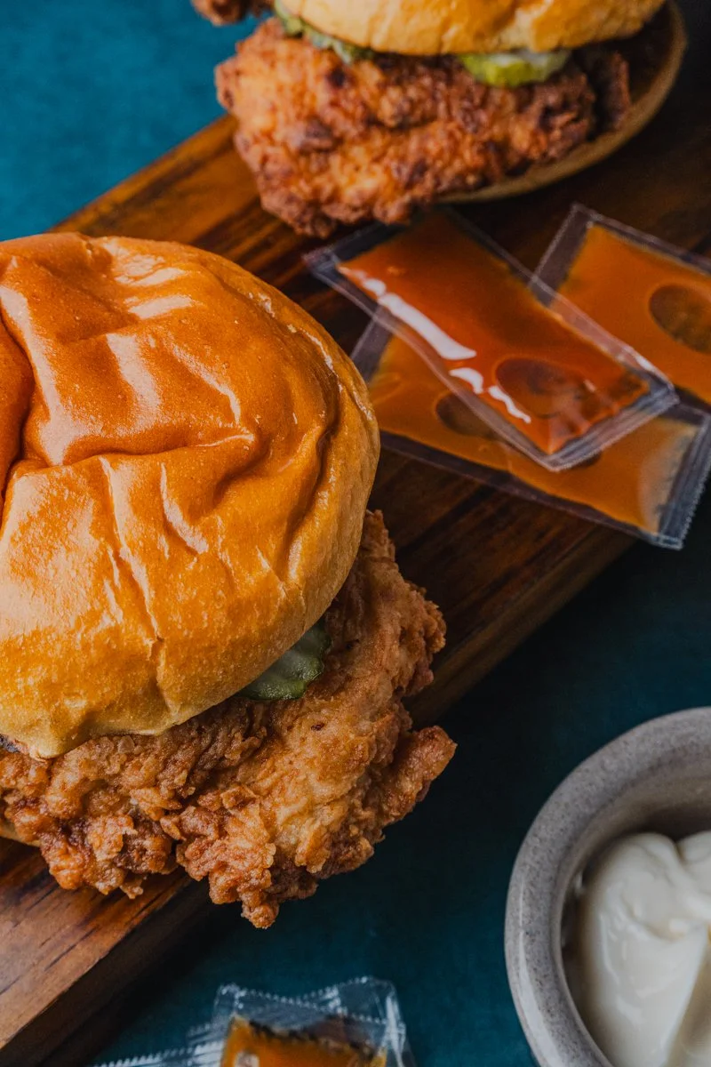 Fried chicken sandwich with a bun, fried chicken fillet, pickles, and sauce, served with packets of hot sauce and mayonnaise.
