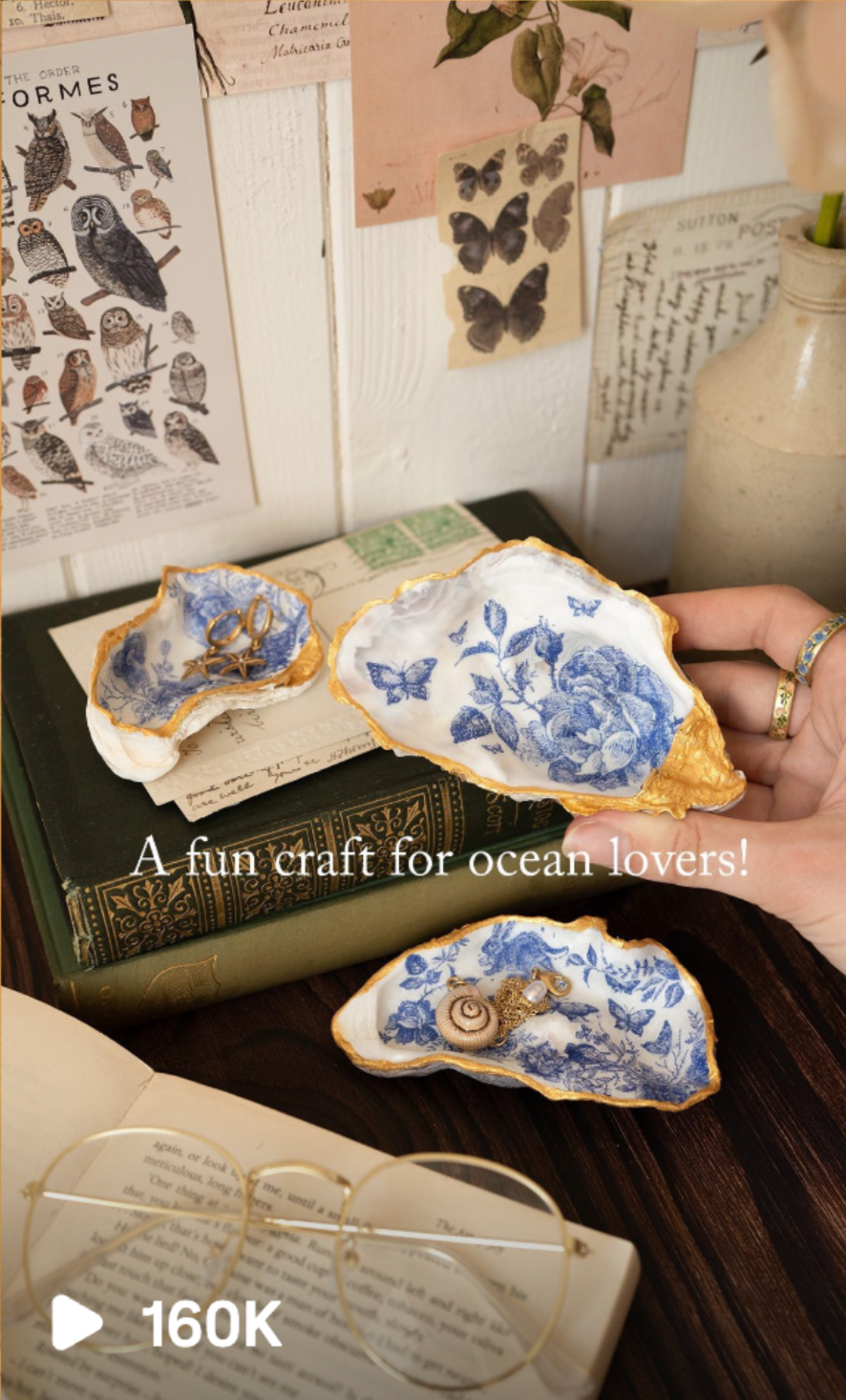 Decorative oyster shells painted with blue floral and butterfly patterns, used as jewelry holders, placed on top of a book and surrounded by vintage decor and botanical prints with butterfly illustrations. Social media manager with content strategy.