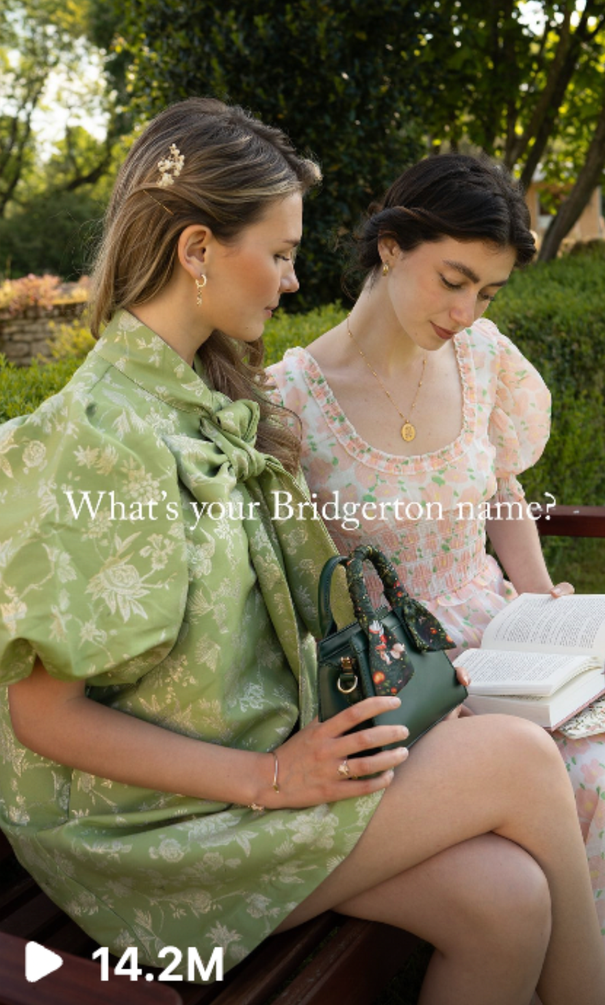 Two women sitting on a park bench, reading a book together, with trees and greenery in the background. Bridgerton cottage core content creators.