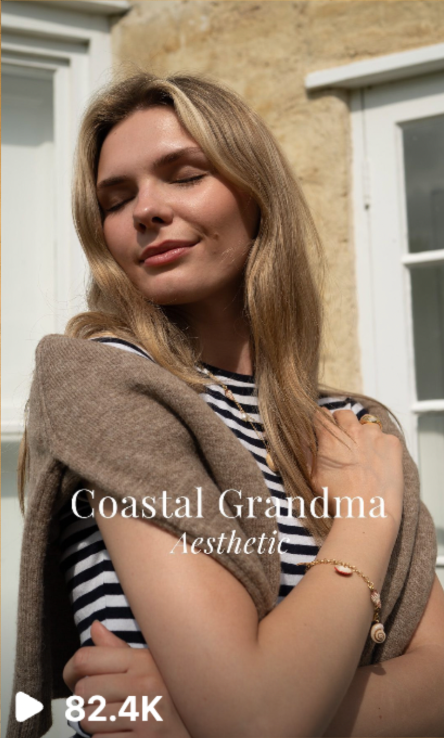 Close-up of a cornish content creator with blonde hair, wearing a striped shirt and a beige sweater draped over her shoulders, standing outdoors near a stone wall and white-framed window. She has her eyes closed, a gentle smile.