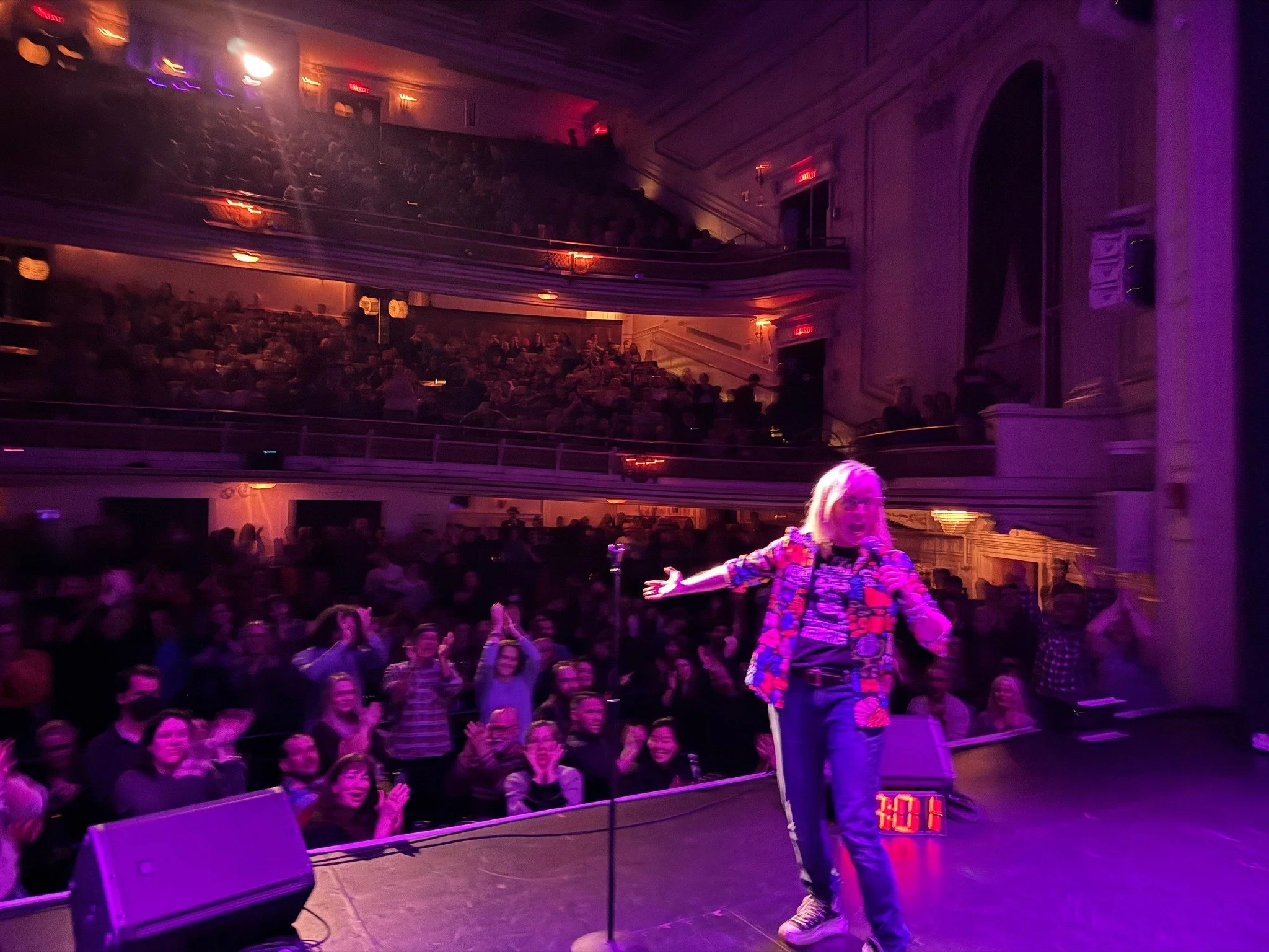 A woman on stage with a microphone facing a large audience in a theater with multiple balconies, illuminated with purple lighting, with a digital clock showing 4:01.