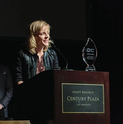 A woman standing at a podium with a glass award on top, at the Hyatt Regency Century Plaza in Los Angeles.